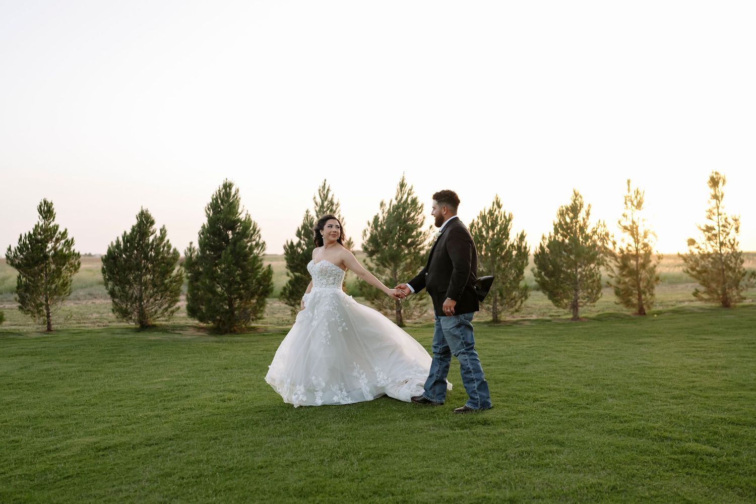 Wide landscape view of a wedding couple embracing on a green lawn with evergreen trees at sunset.