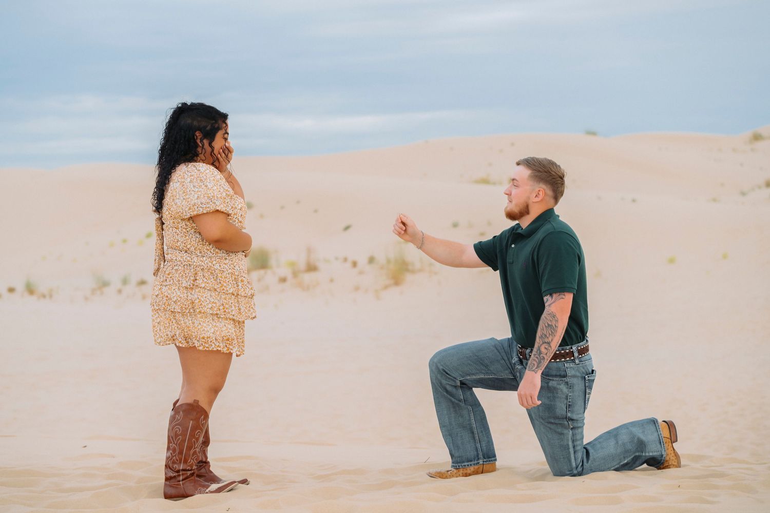 A couple in casual attire poses for photos during a romantic desert proposal at sunset.