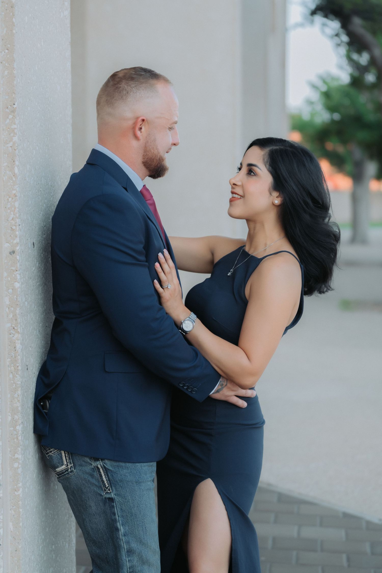 Romantic couple poses together against white column outdoors, with woman in dark formal dress and man in navy blazer for their engagement