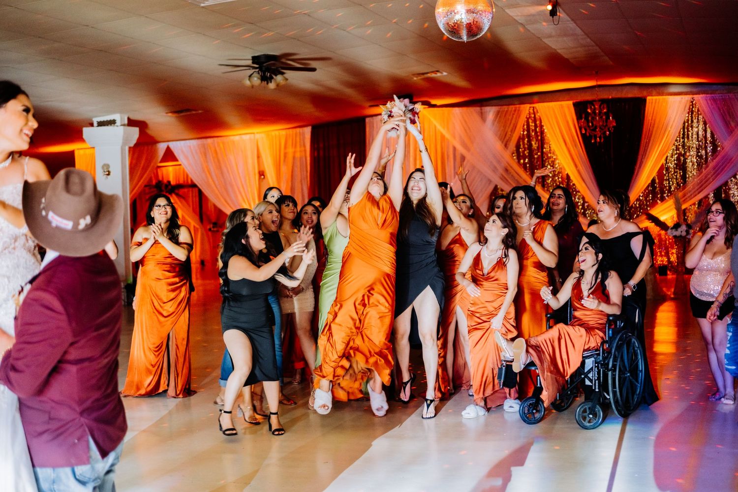 Large group dancing in formation at wedding reception with orange ambient lighting.
