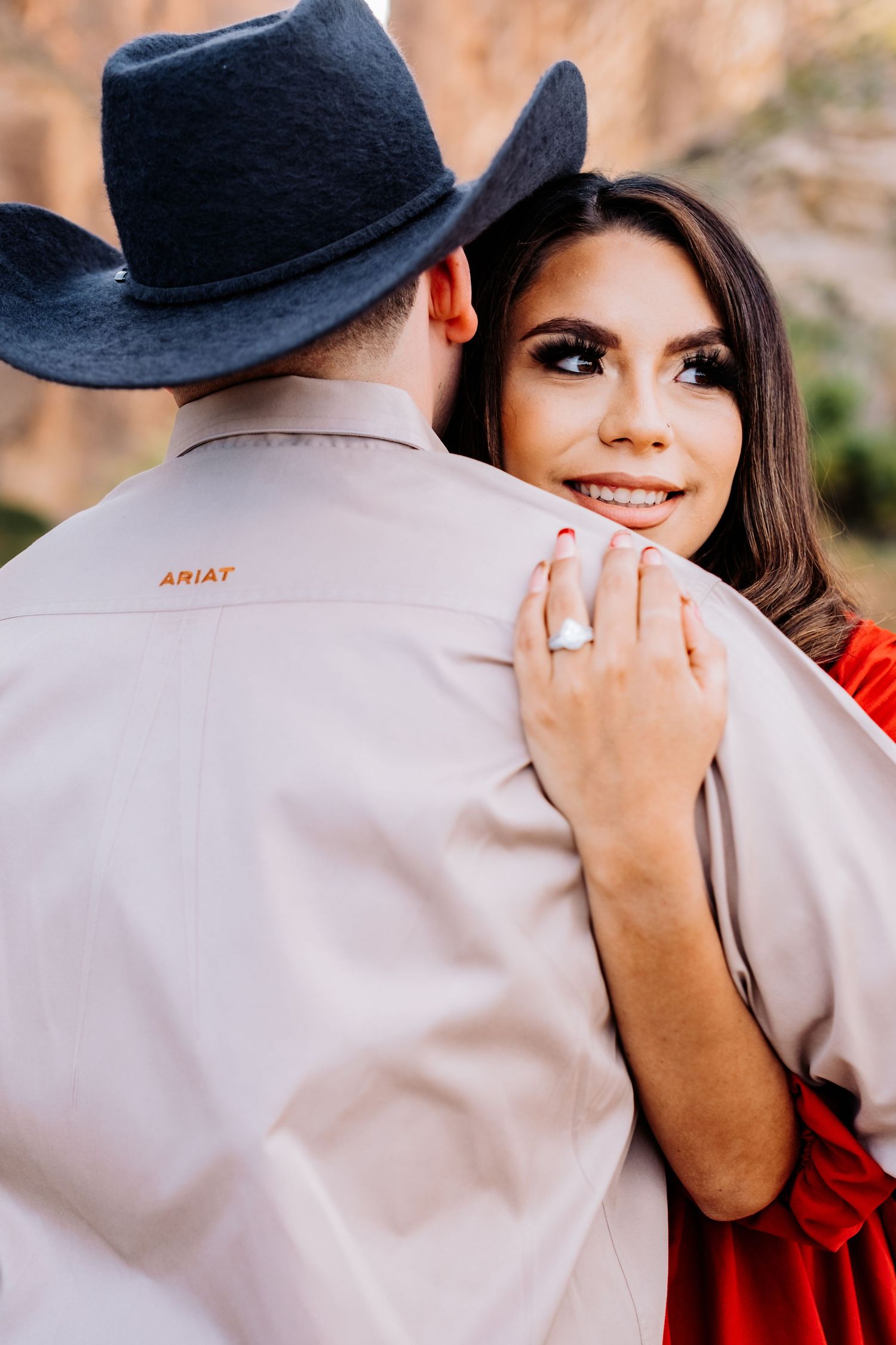 Couple embracing near canyon walls with woman in red dress showing engagement ring.