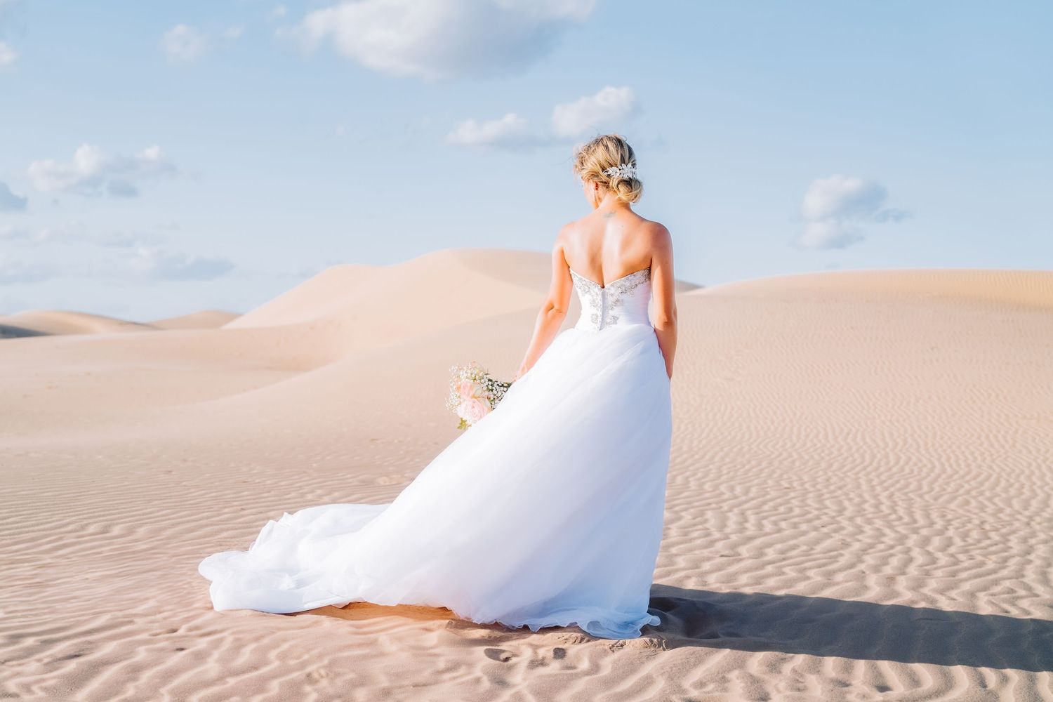 Back view of bride in flowing white gown standing on desert sand dune.