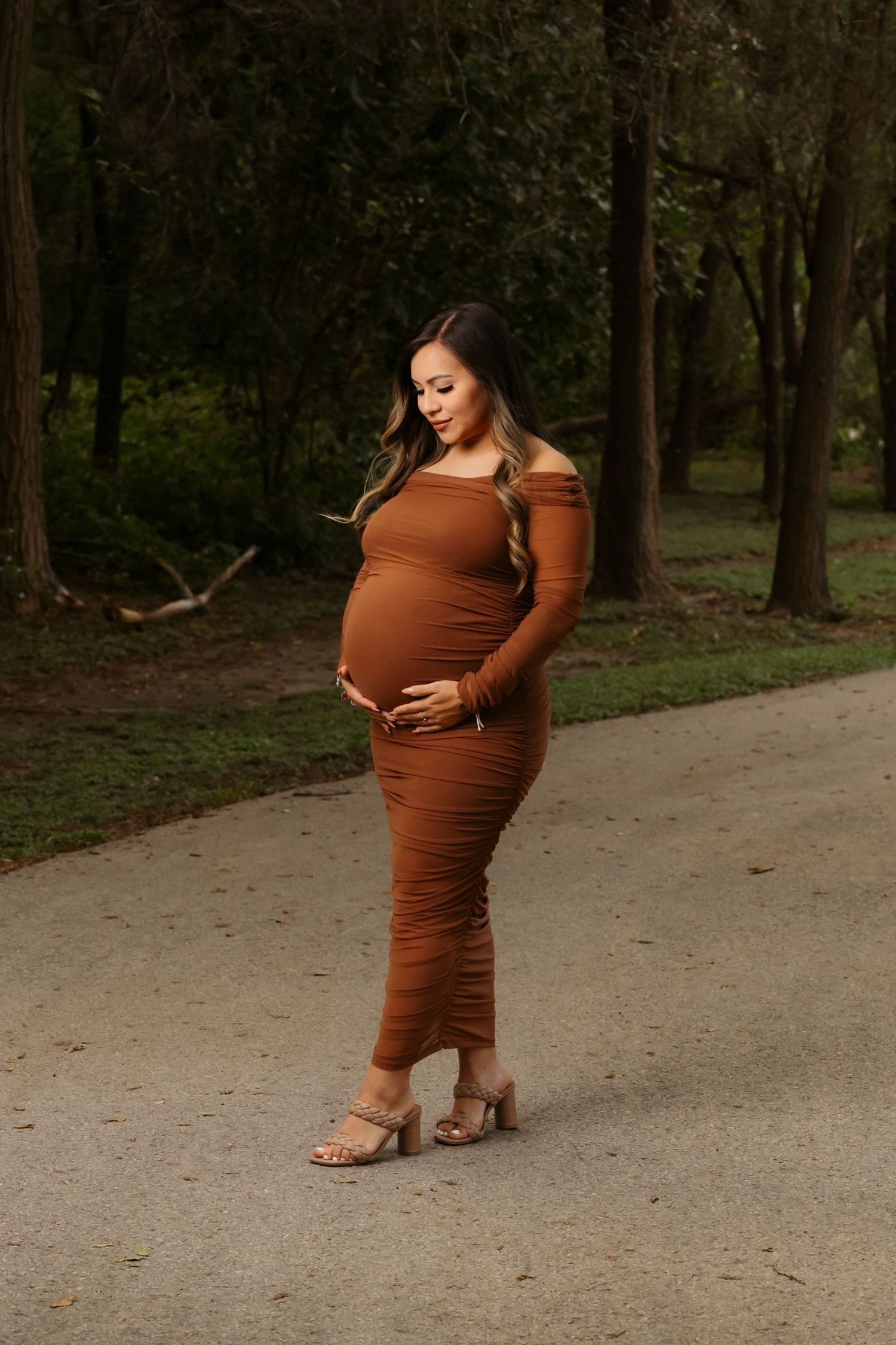 A woman in a rust-colored dress cradles her baby bump while standing on a dirt path surrounded by trees.