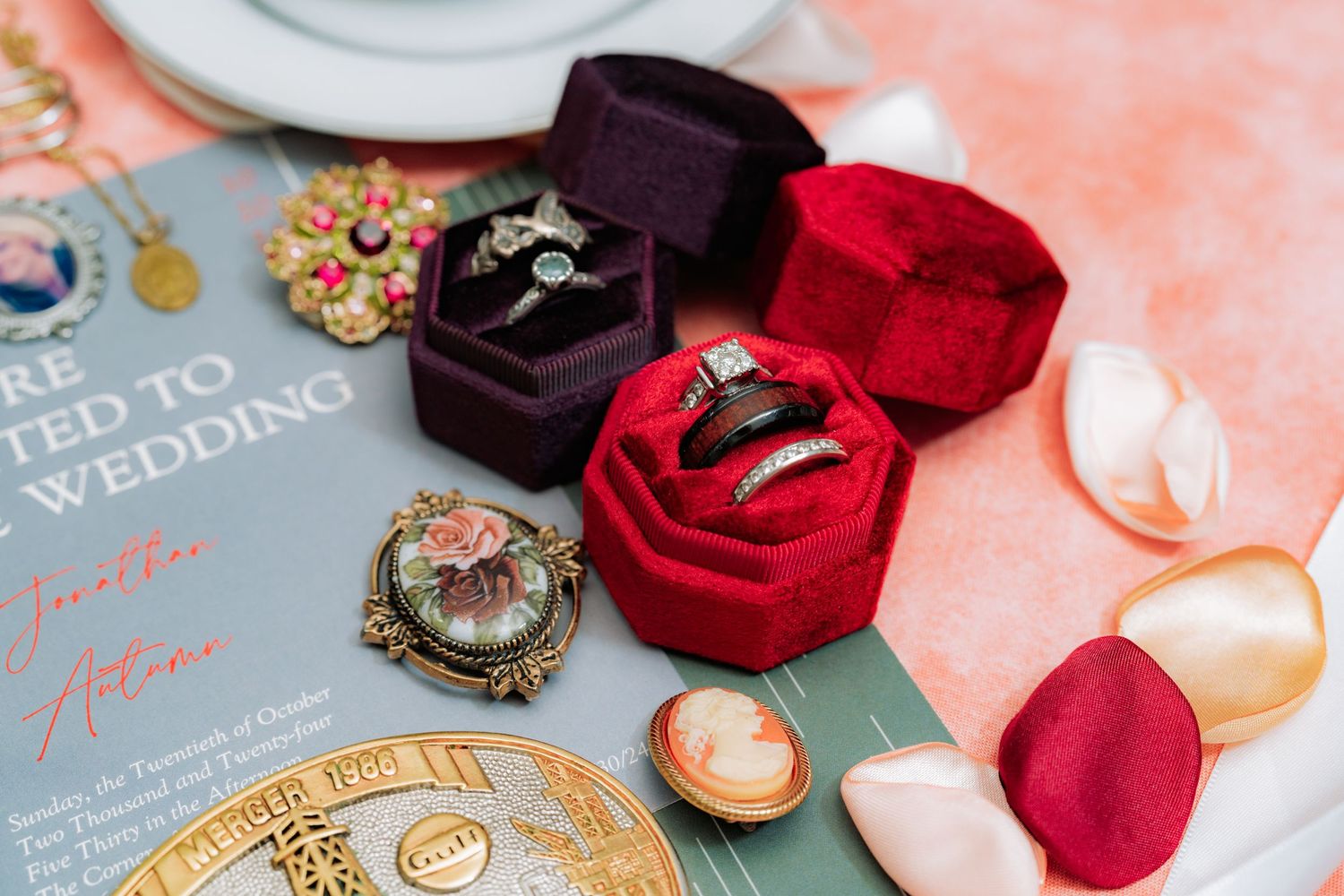 Elegant wedding accessories including ring boxes, red roses and invitation details on coral backdrop.