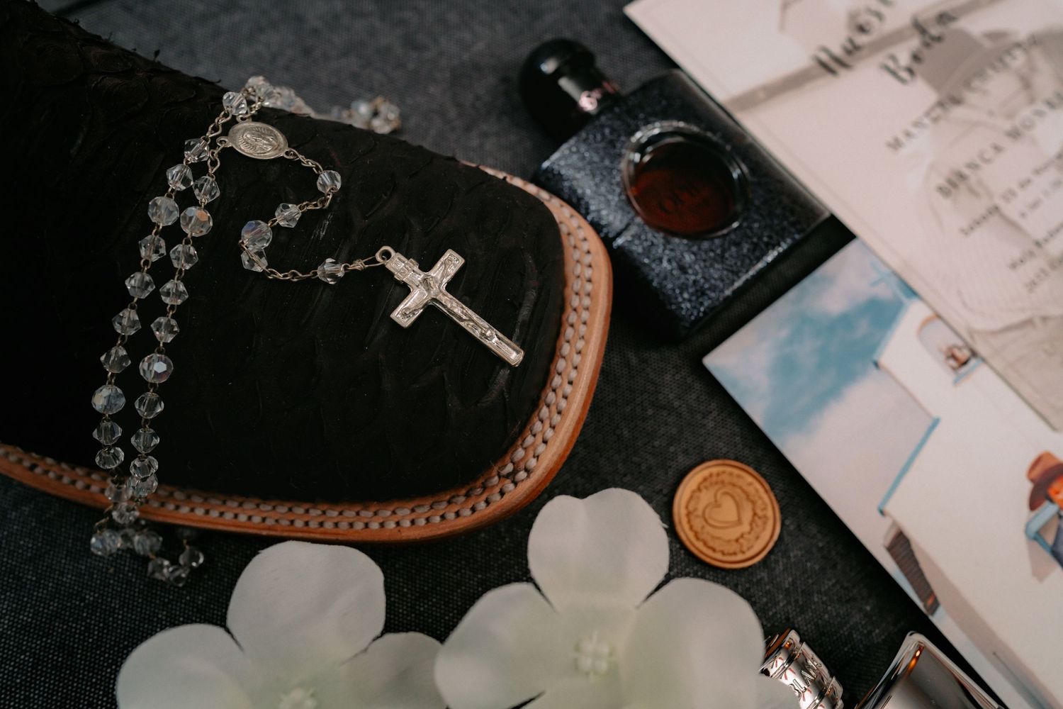 Elegant wedding stationery and accessories arranged on black surface with rosary and ribbon details.
