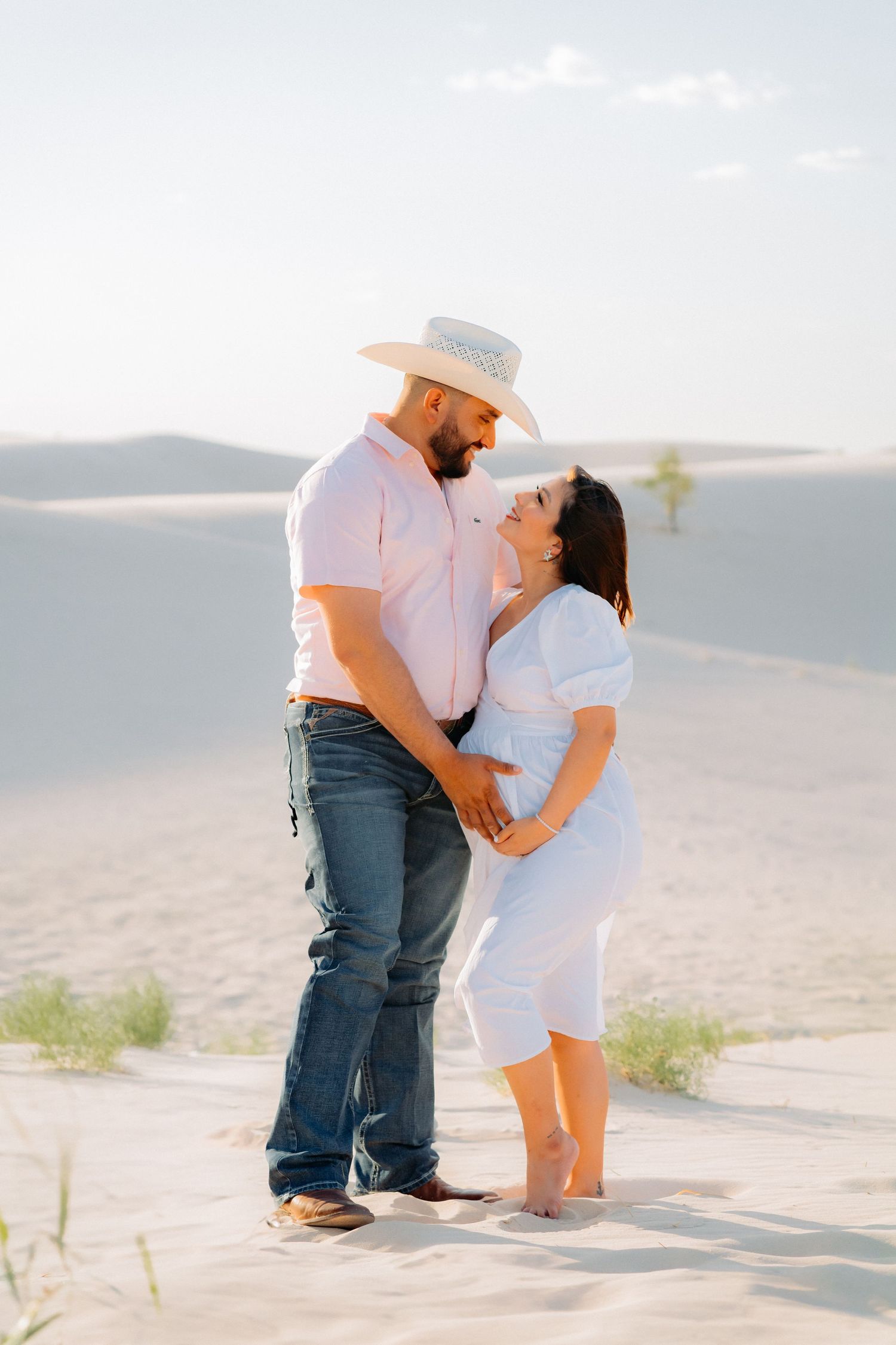 A couple embraces in white outfits against a scenic desert landscape during sunset.