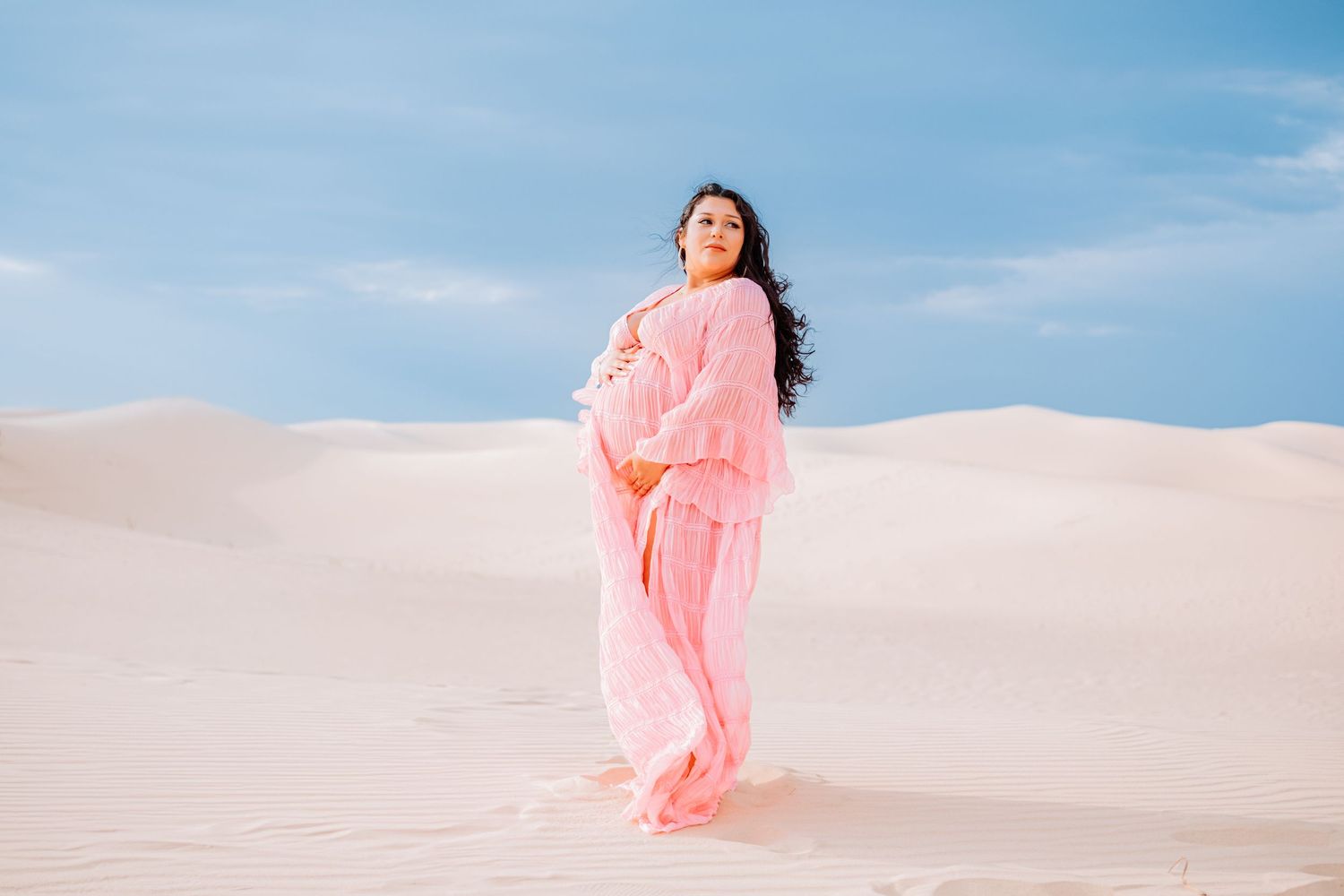 Woman in flowing pink dress walking across white sand dunes under blue sky.