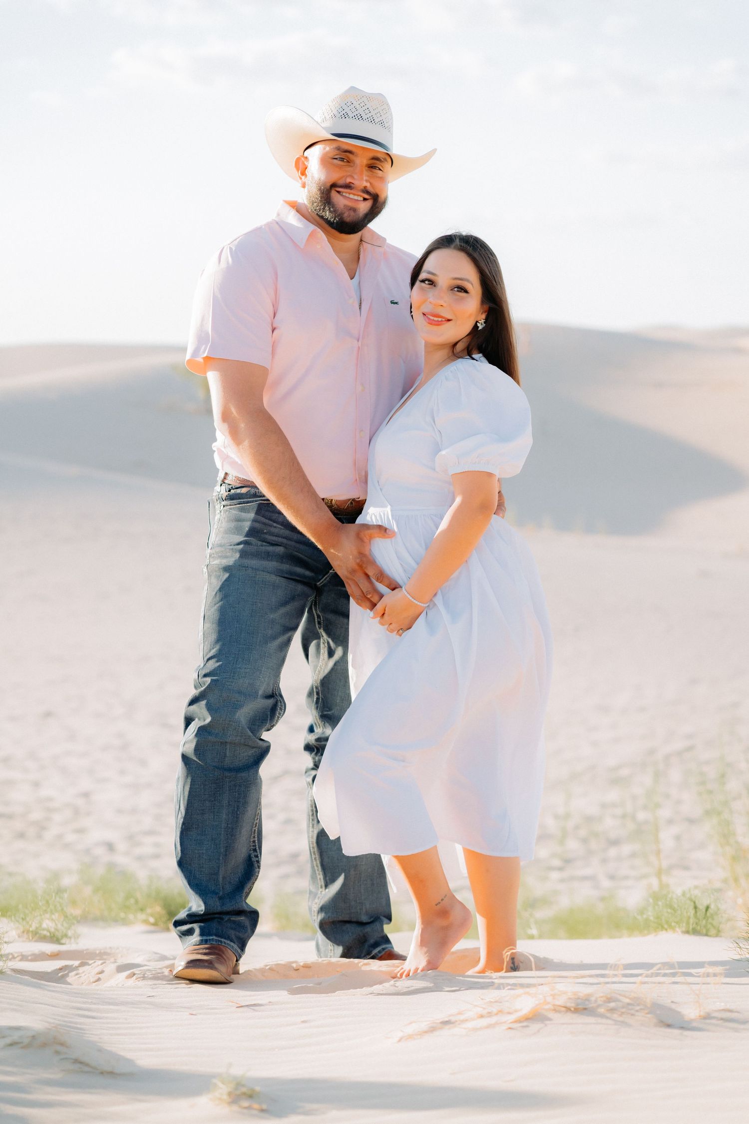 Western-themed maternity shoot featuring couple in white dress and cowboy hat on desert dunes.