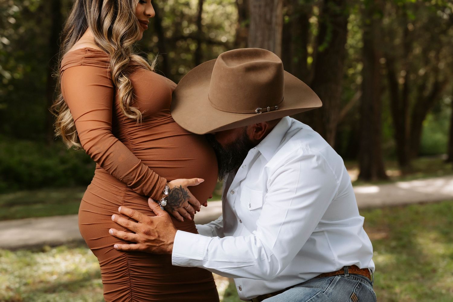 A couple in brown and white outfits embraces tenderly during a romantic outdoor photoshoot in a wooded setting.