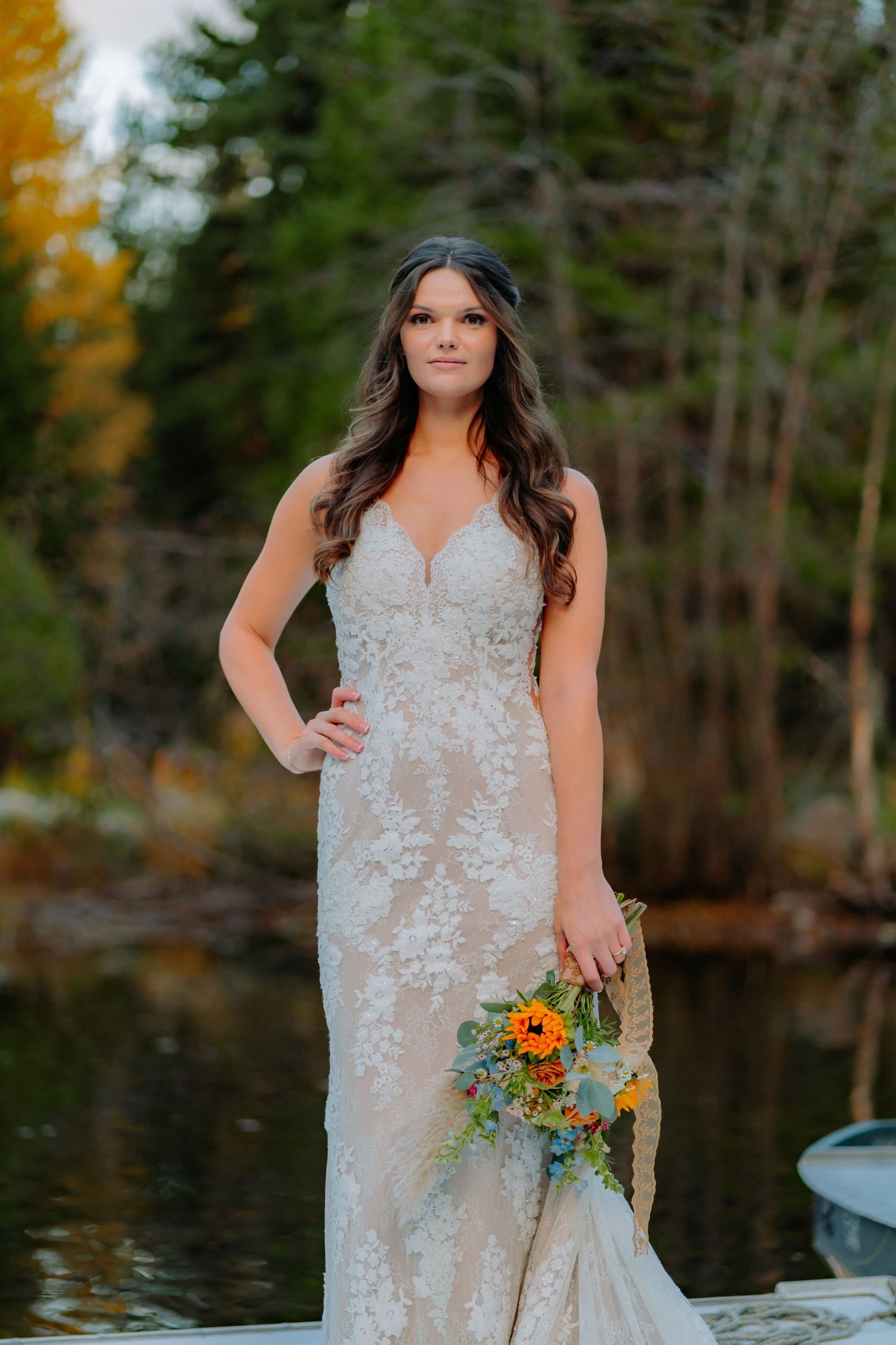 Portrait of person in fitted lace wedding gown holding sunflower bouquet on dock with lake backdrop.