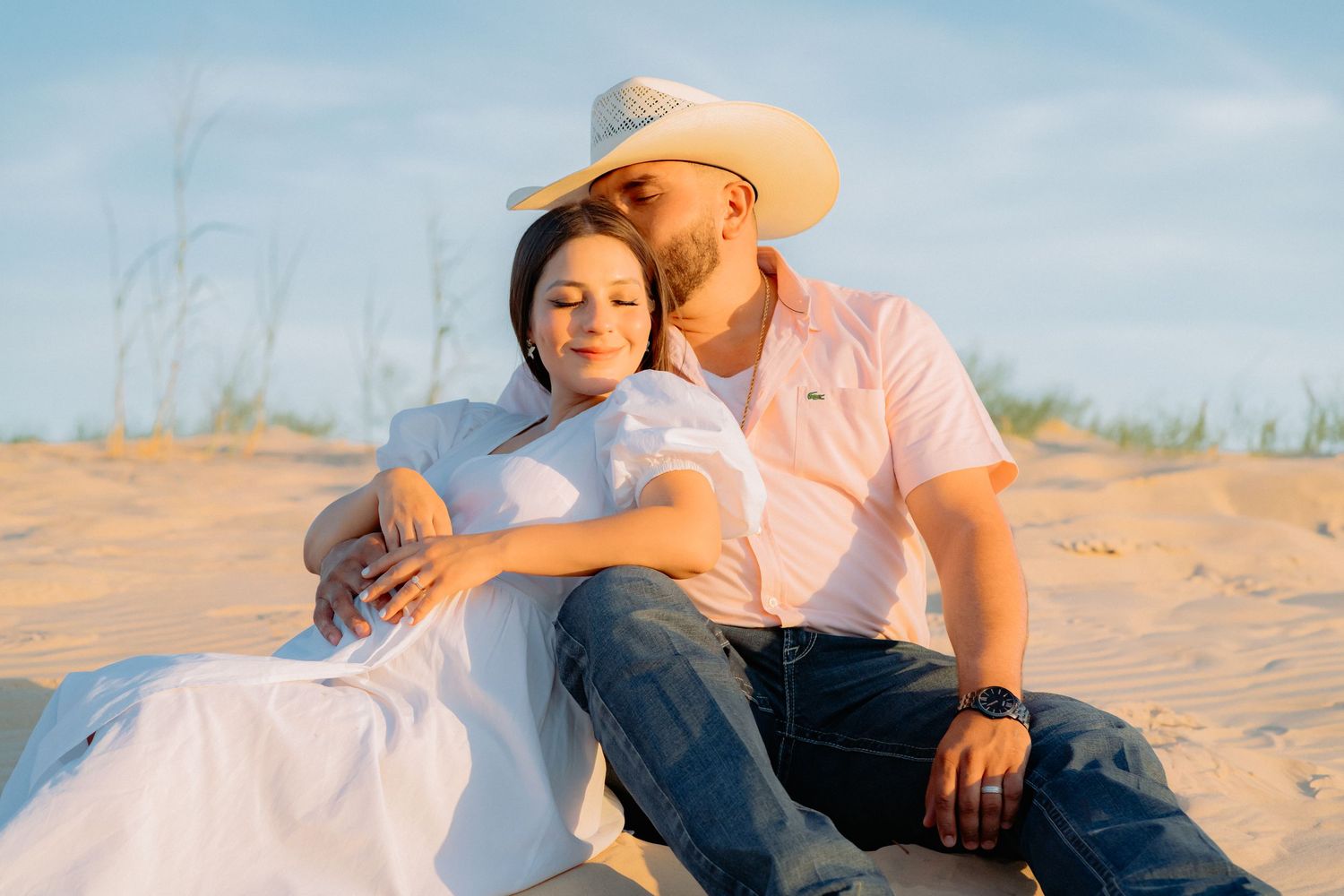 Romantic maternity photo of couple sitting together on sand dunes at sunset.