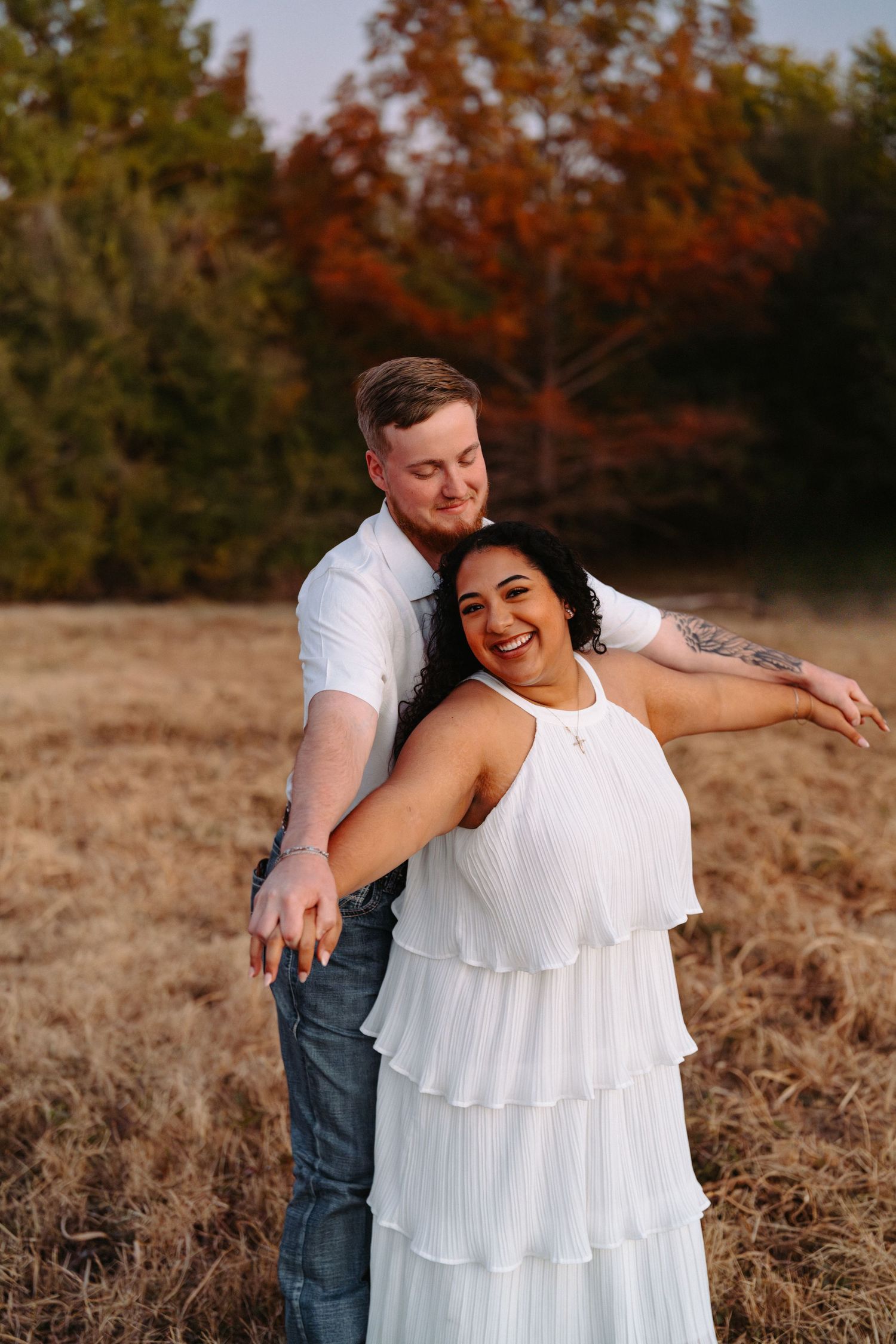 A couple embraces in a field during autumn with fall foliage visible in the background.