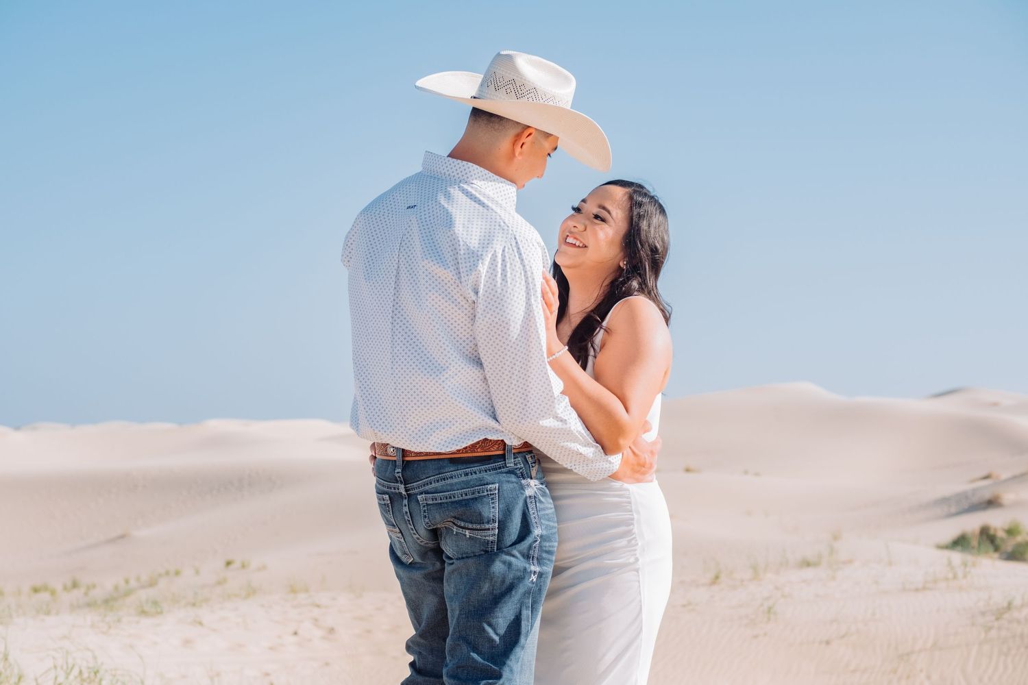 Couple embracing on sand dunes against bright blue sky.