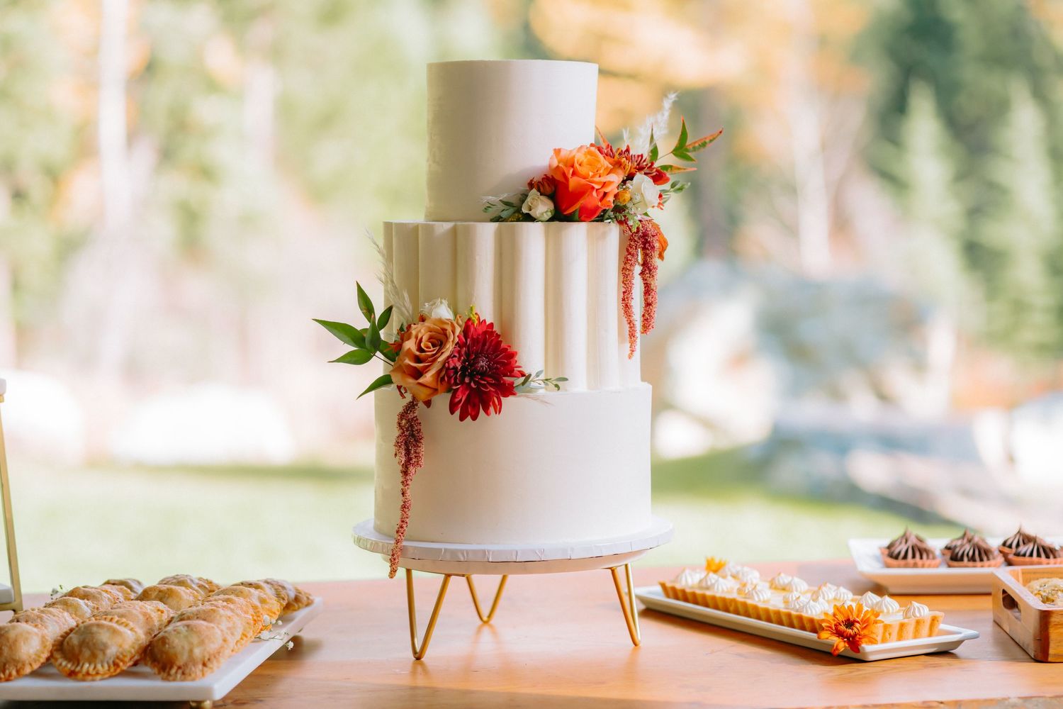 Elegant white tiered wedding cake decorated with coral flowers and greenery displayed on a gold stand with desserts nearby.