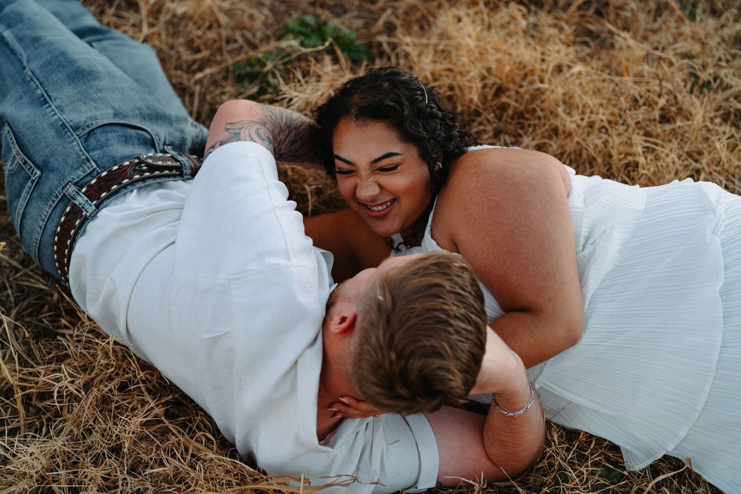 A couple in white outfits laying in golden grass sharing an intimate moment together during a photoshoot.