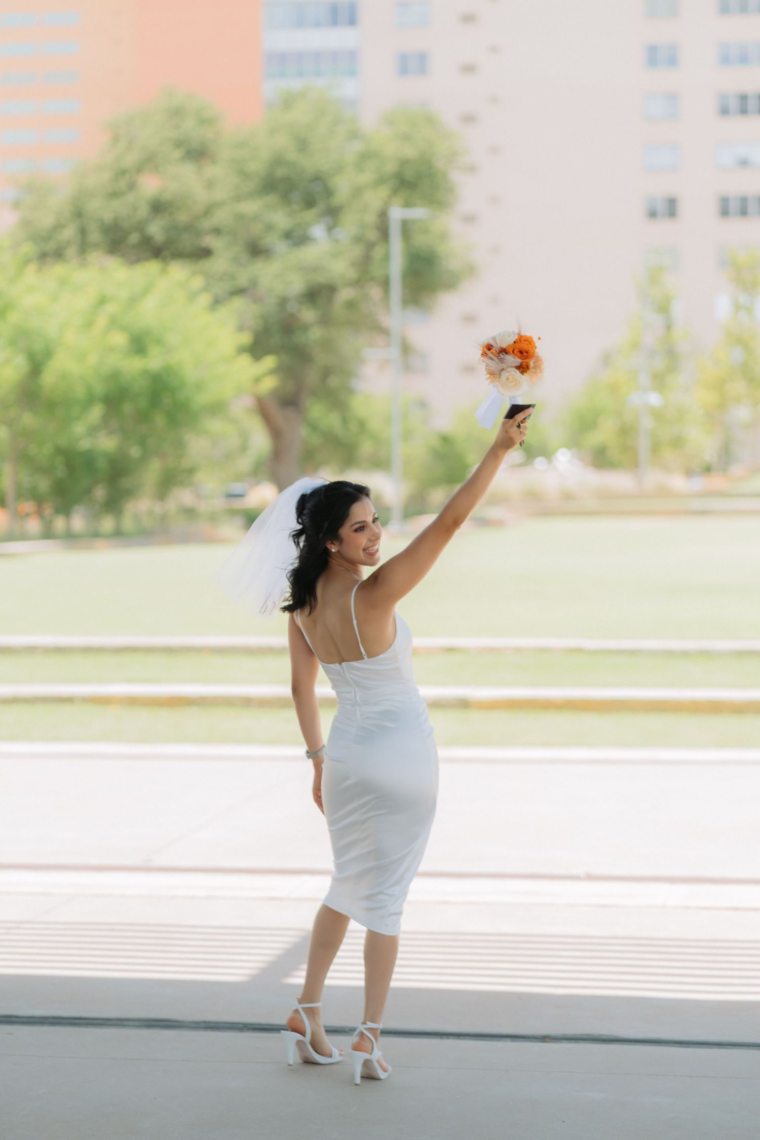 A bride in a fitted white dress poses joyfully with raised arms holding orange flowers outdoors.