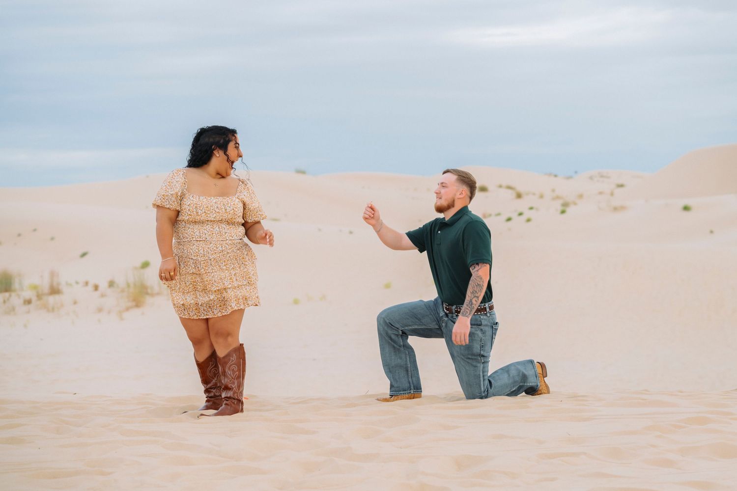 A couple shares a romantic proposal moment on sandy dunes during golden hour at the beach.
