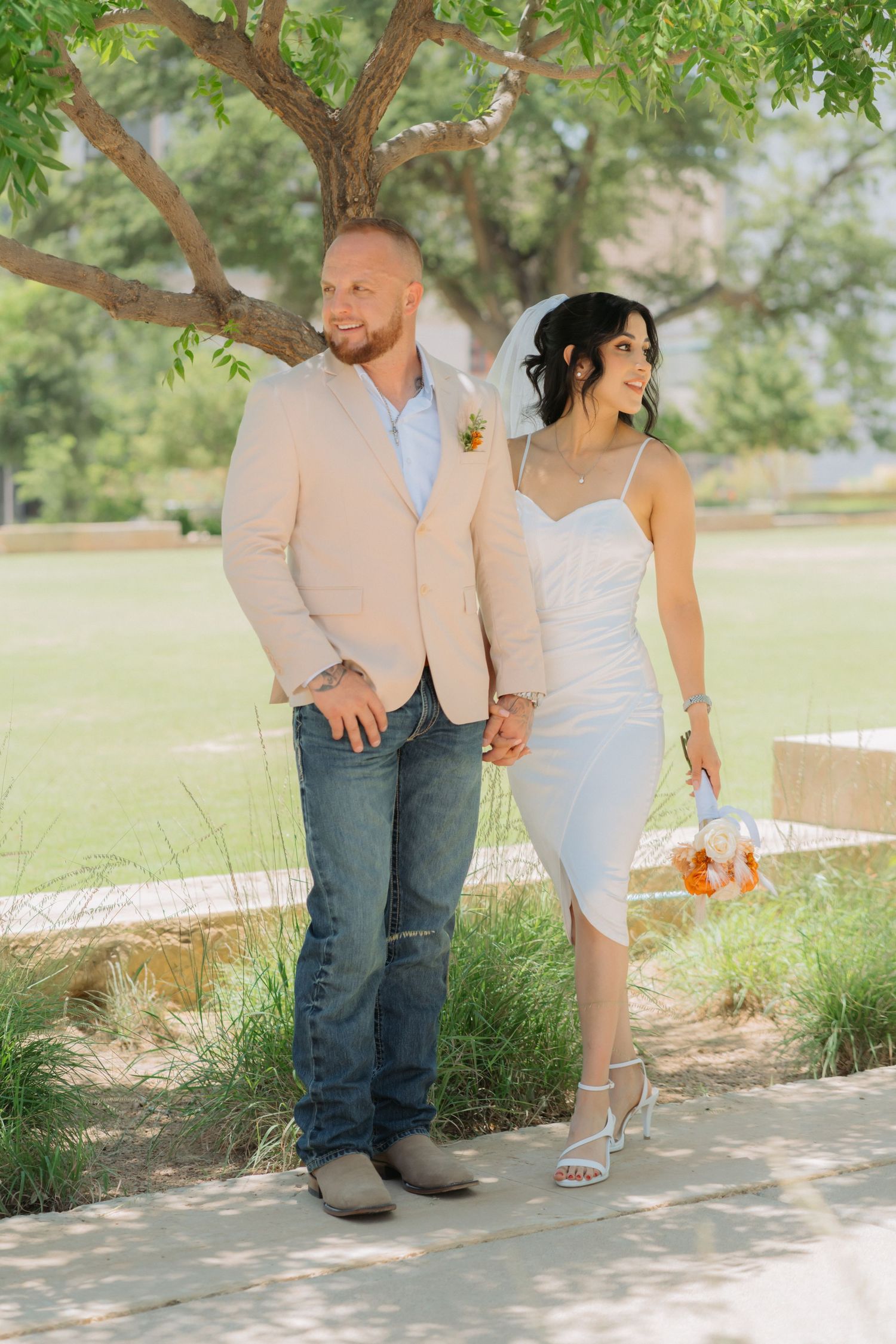 A couple in casual attire poses along a garden path with jeans, boots and a white sundress in a romantic summer setting.