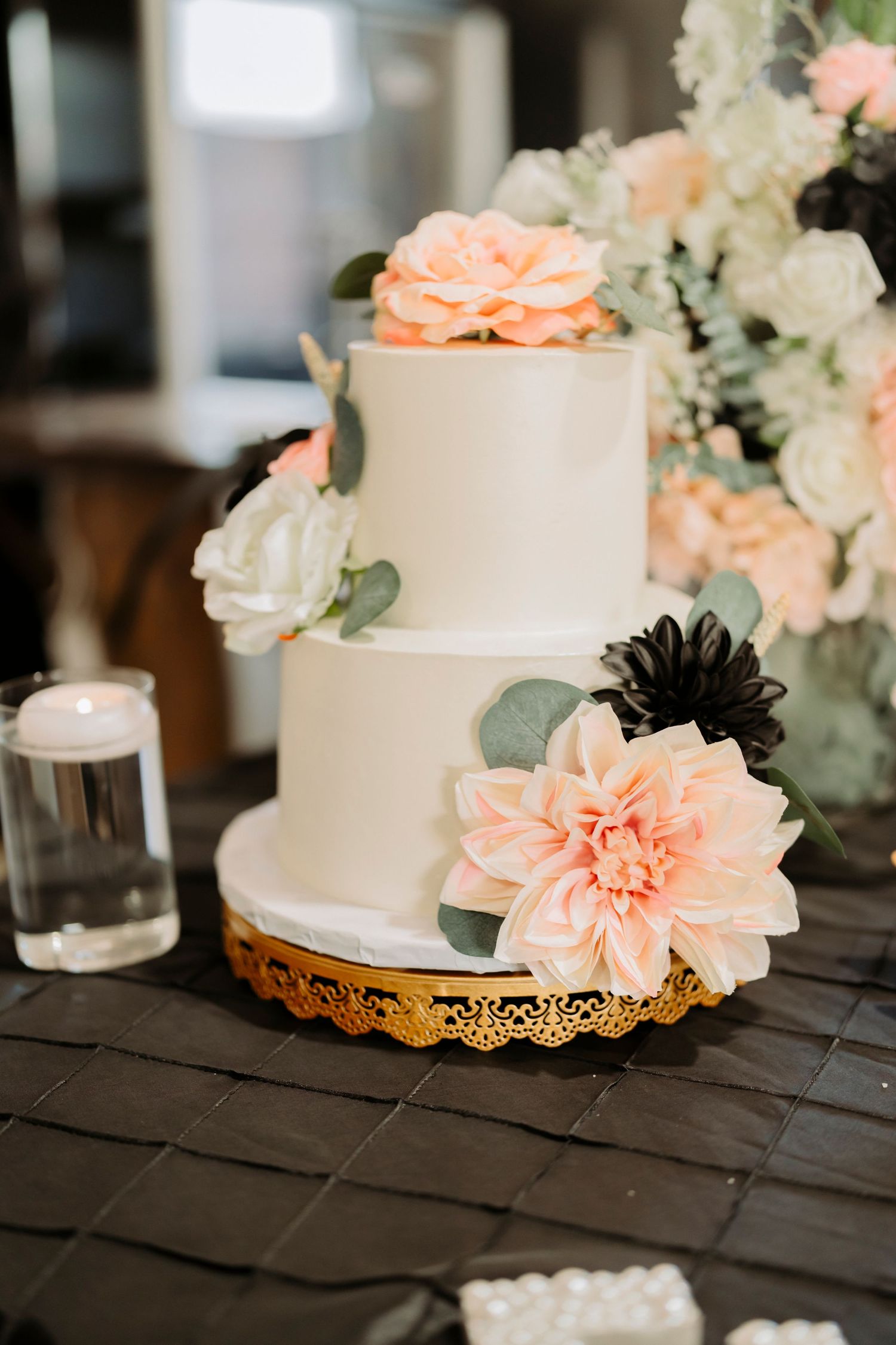 Elegant two-tier white wedding cake decorated with peach and black flowers on gold stand.