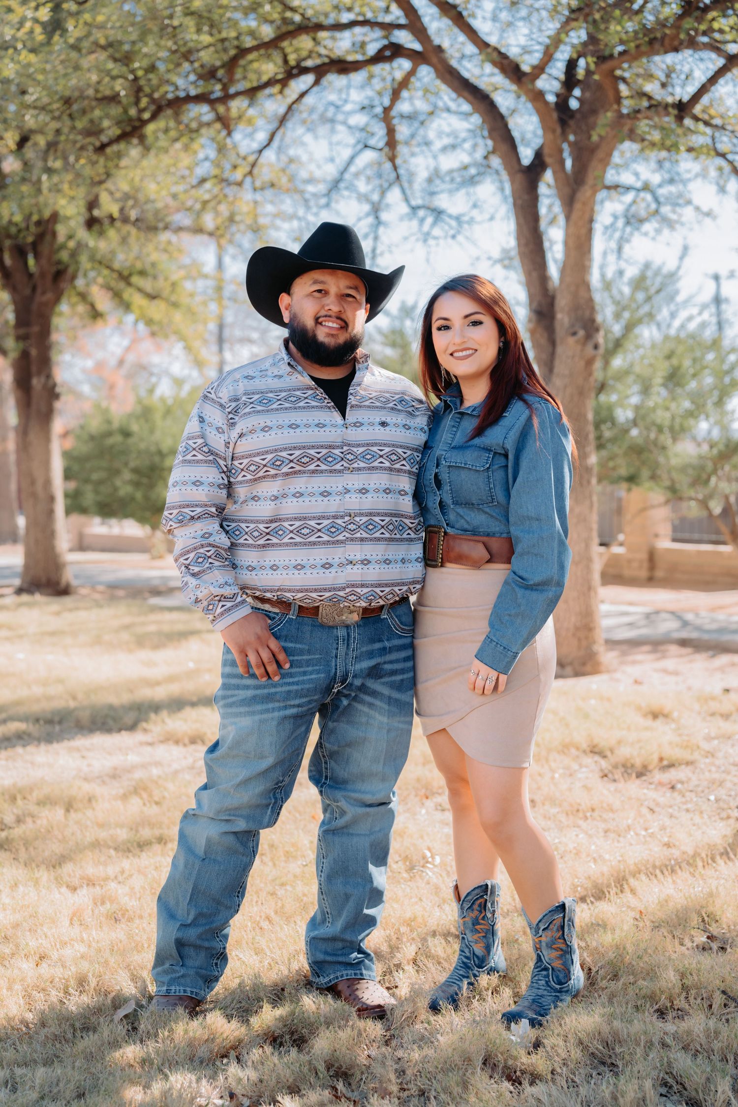 Couple poses together under trees in casual western attire during autumn photoshoot.