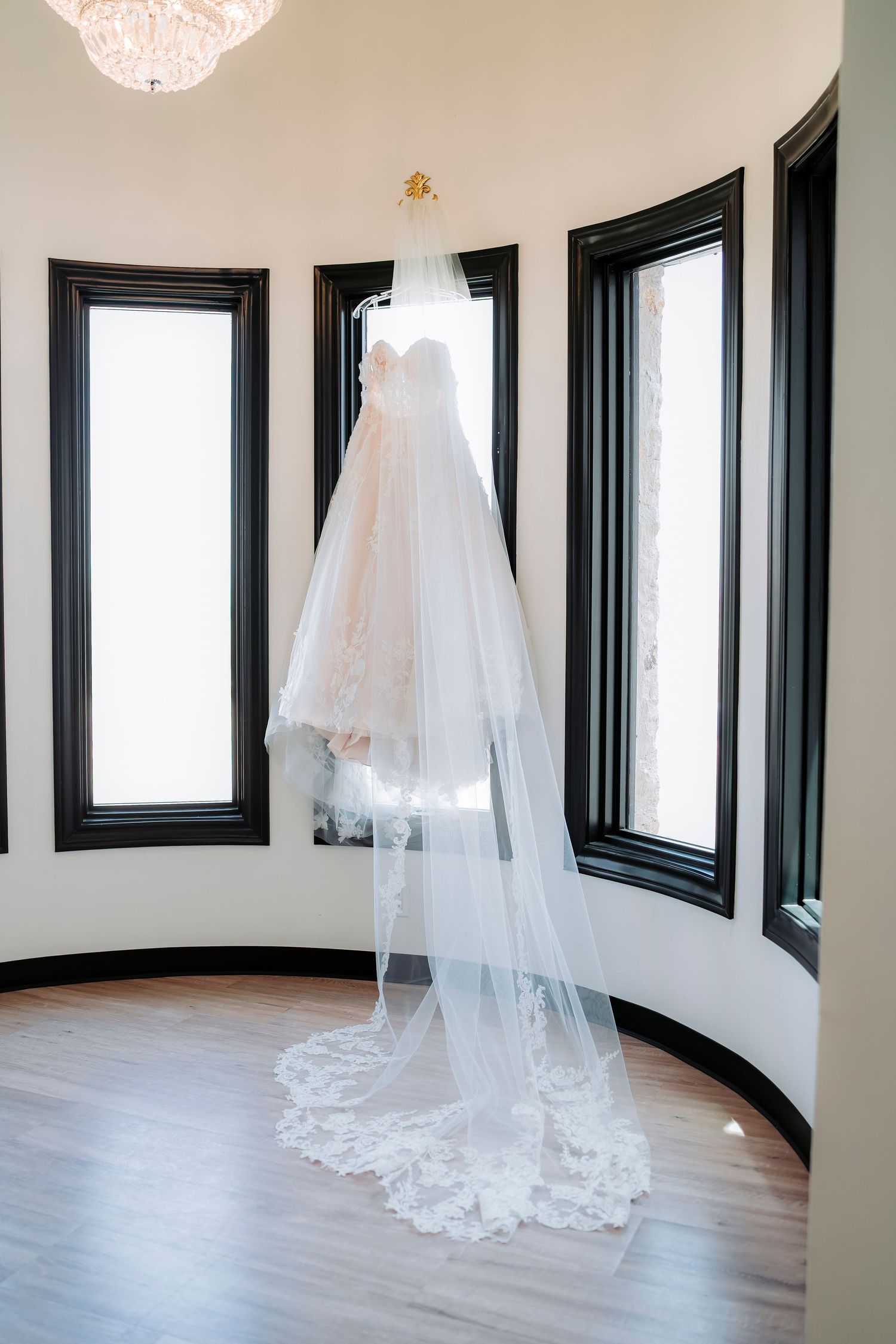 A white wedding dress and long lace veil hang elegantly in a curved room with dark-trimmed windows and hardwood floors.