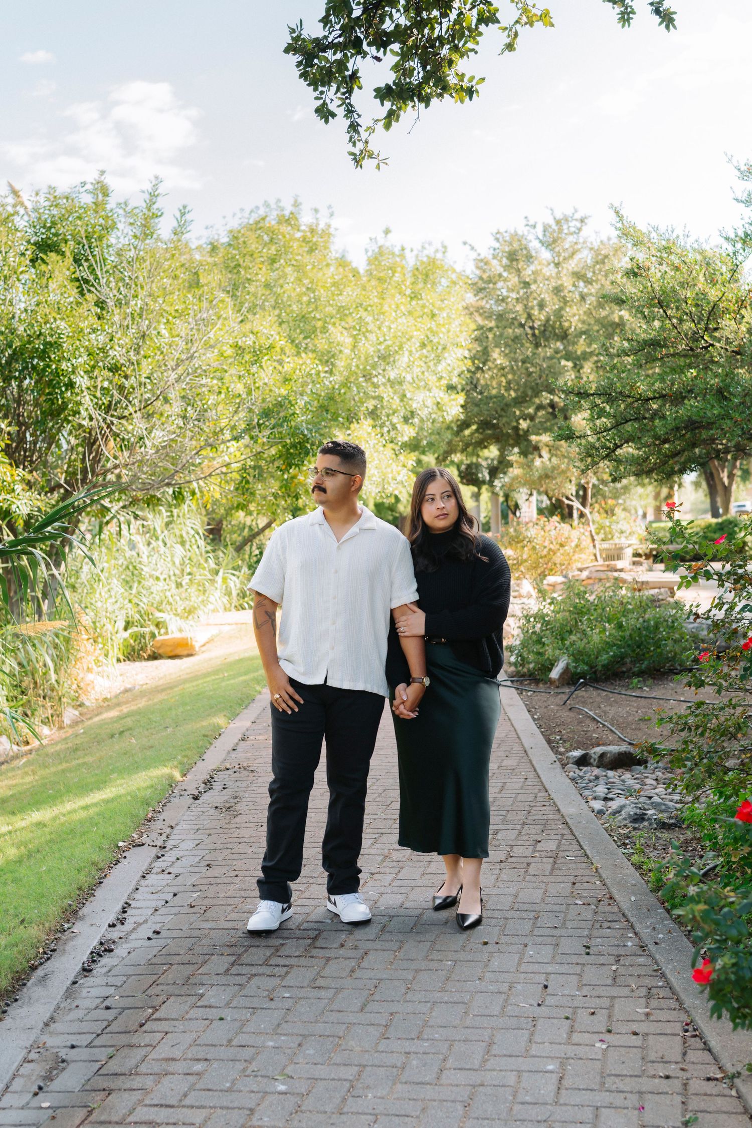 Three people walk together on a brick pathway surrounded by landscaped gardens on a sunny day.