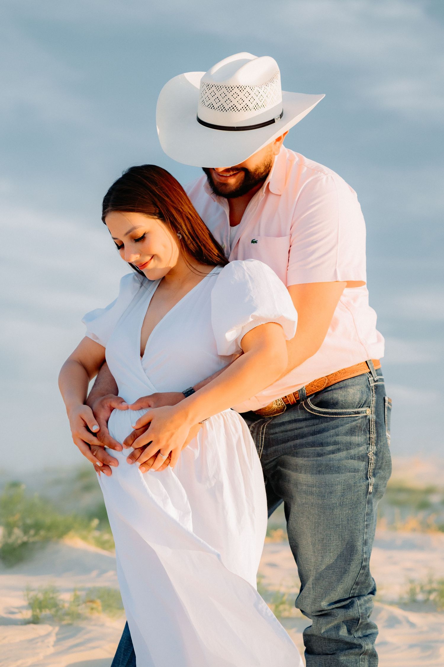 Couple in white outfits embrace during maternity photoshoot with man wearing cowboy hat.