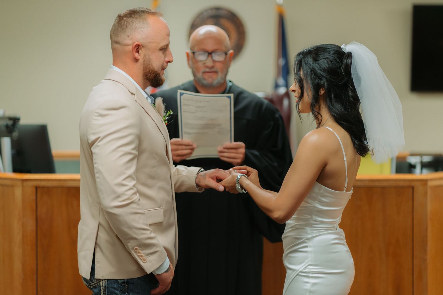 A loving couple exchanges rings during an intimate courthouse wedding ceremony while the officiant reads from documents.