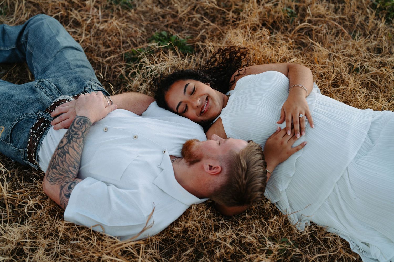 Couple in white clothing lying together on dry grass during golden hour photography session.