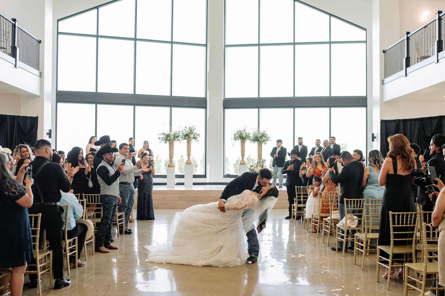 A couple shares their first kiss as newlyweds in a modern venue with floor-to-ceiling windows while guests capture the moment.