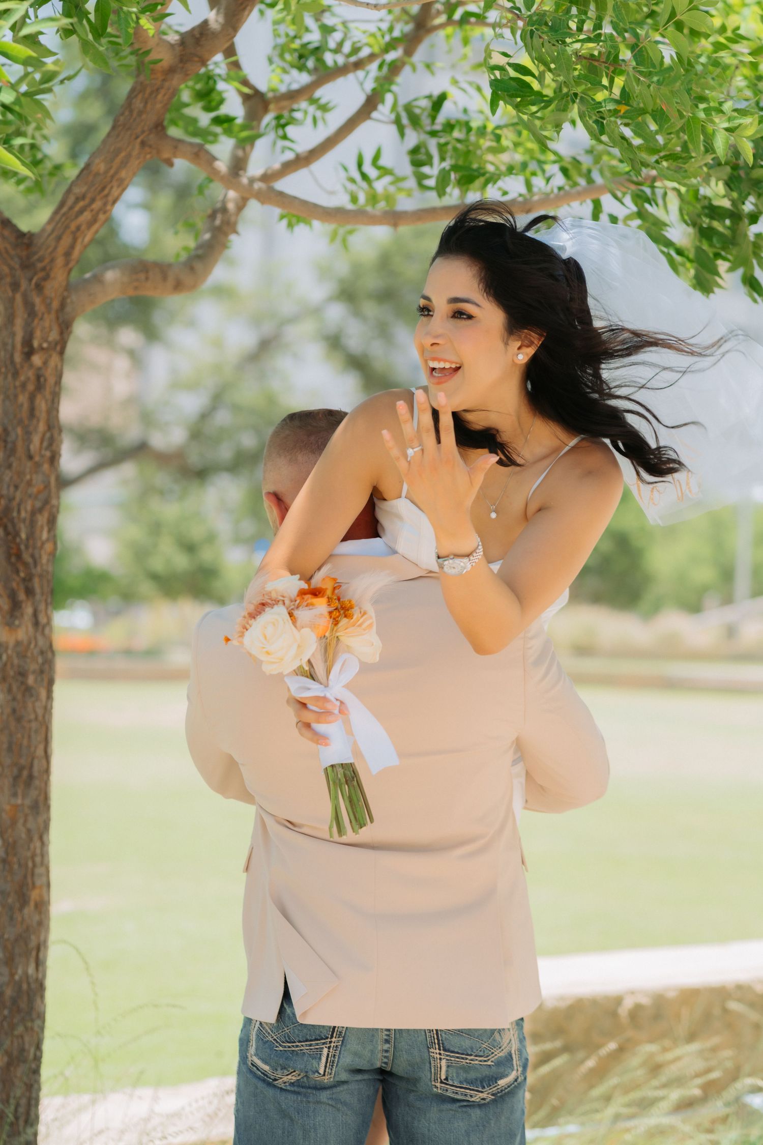 Person in a beige sweater stands in front of trees while wind blows through their dark hair and clothing.