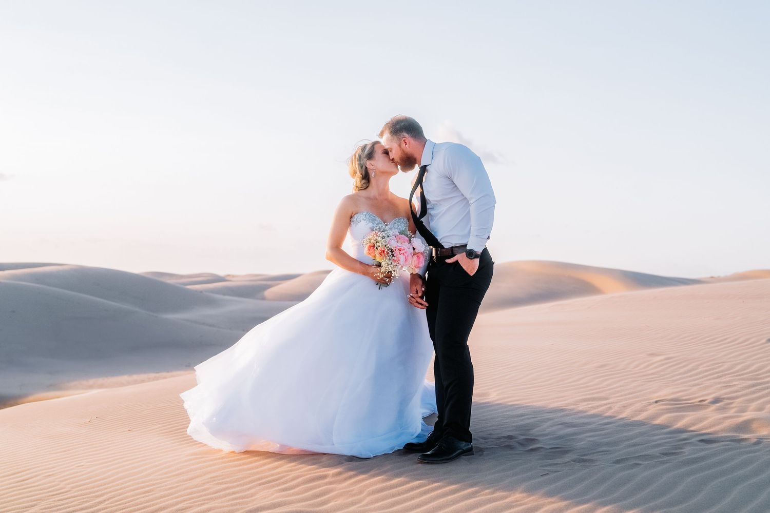 Elegant wedding photography in desert landscape with billowing white dress against sand dunes.