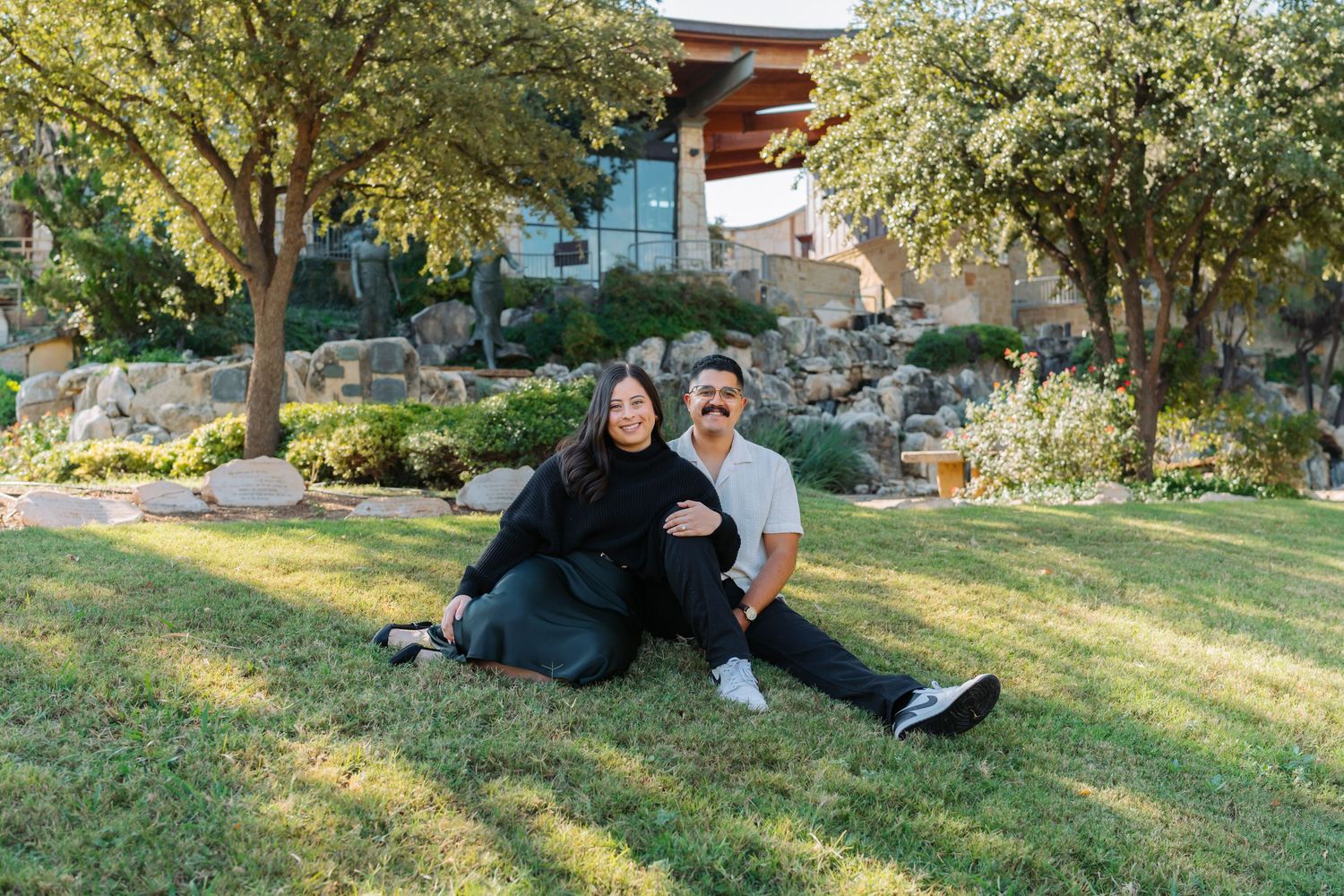 A couple relaxes together on a grassy hillside during a sunny afternoon in a park setting.