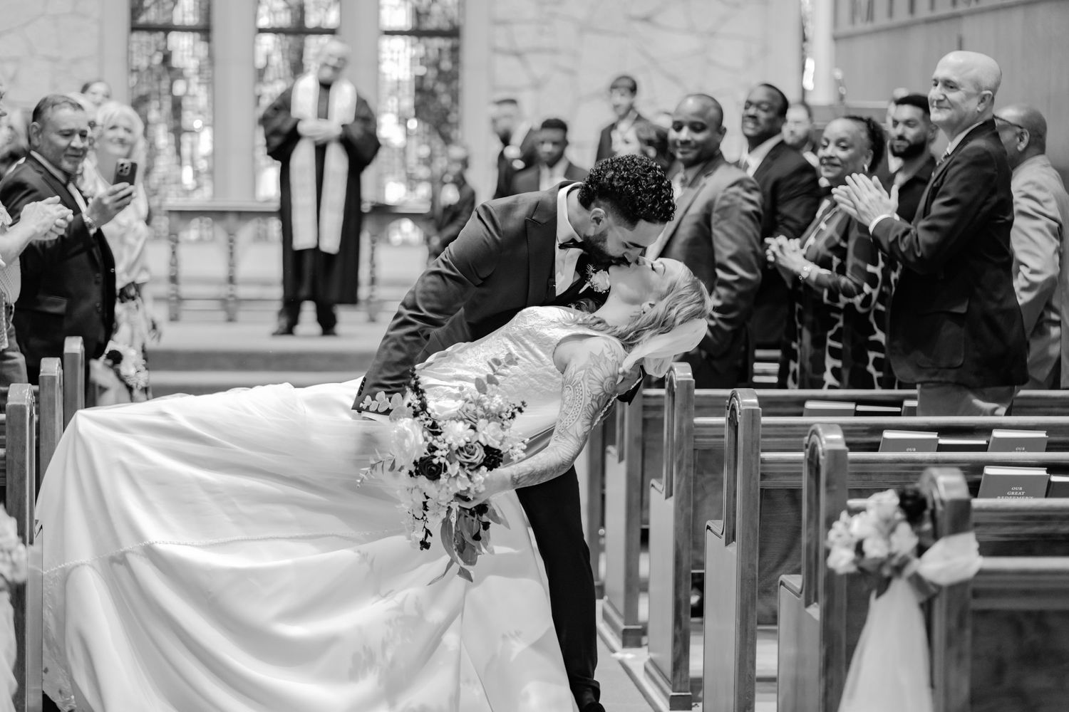 Black and white wedding photo showing guests reacting joyfully to couple's romantic moment in church.