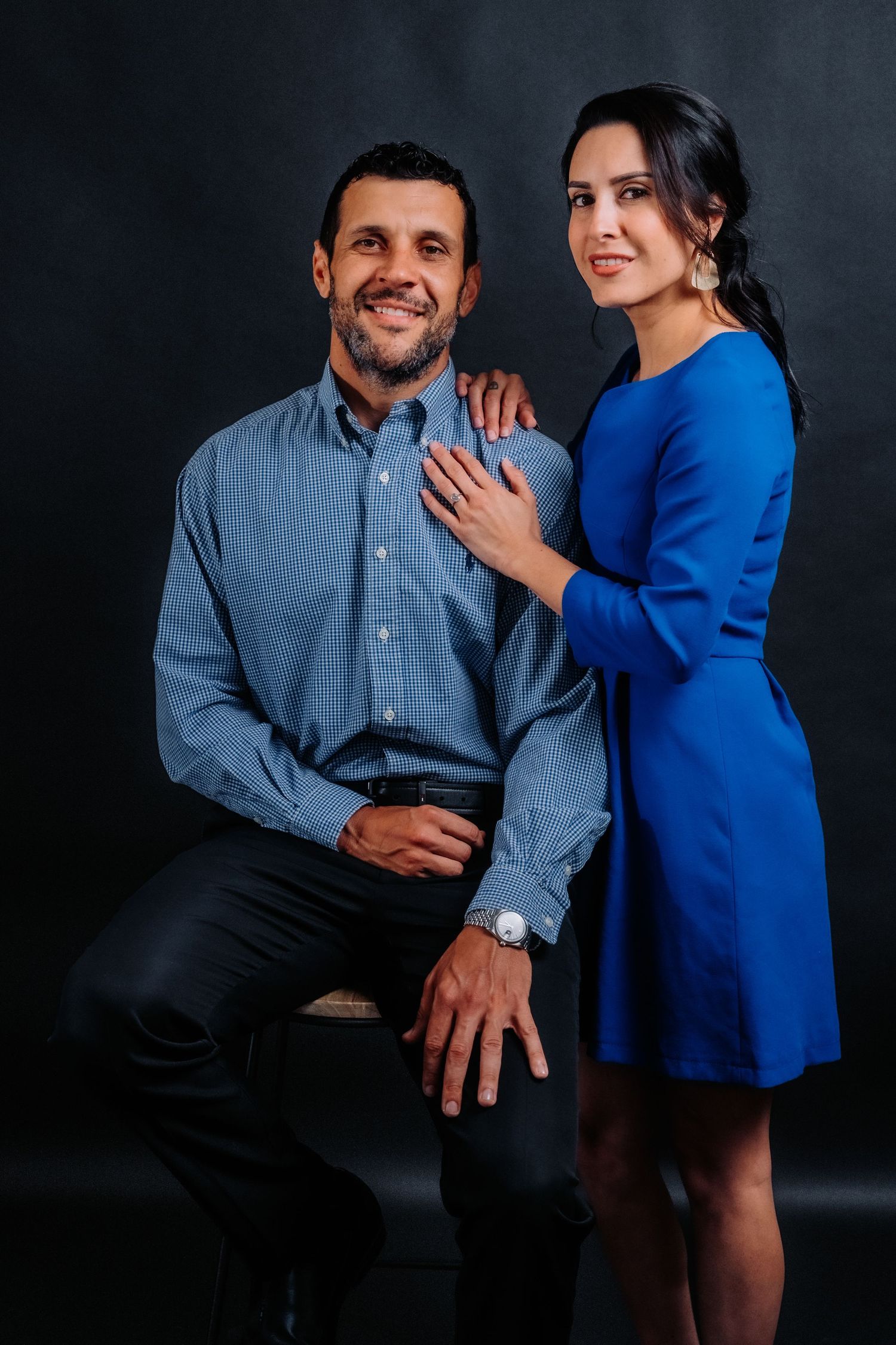 Engagement photo of couple in coordinating blue outfits against dark studio background.
