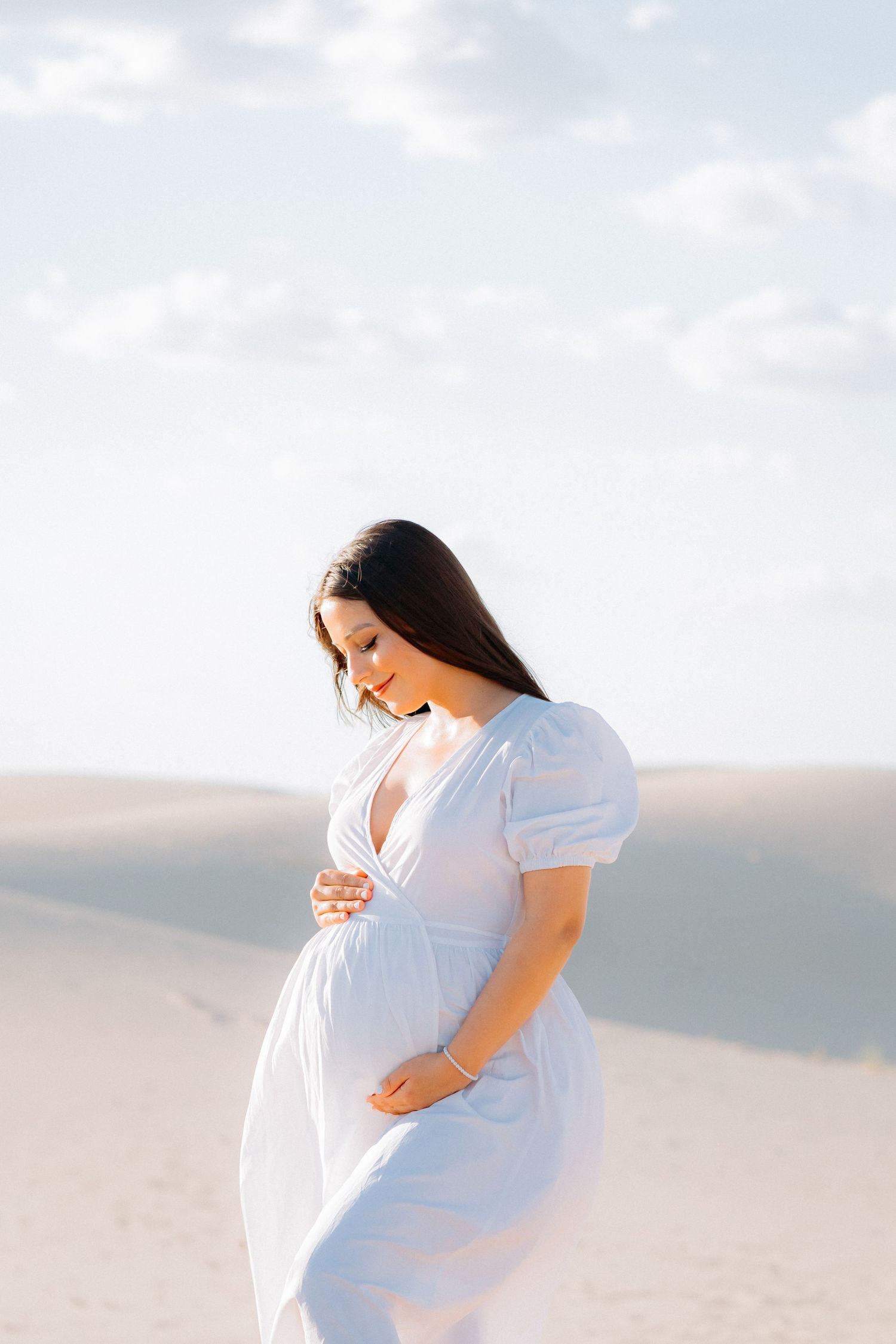 Side view of expectant mother in white dress touching belly against desert landscape.