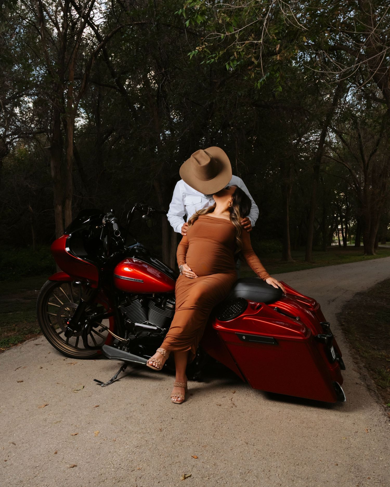 A person in brown pants and cowboy hat poses with a custom red Harley-Davidson motorcycle on a dirt path in a wooded area.