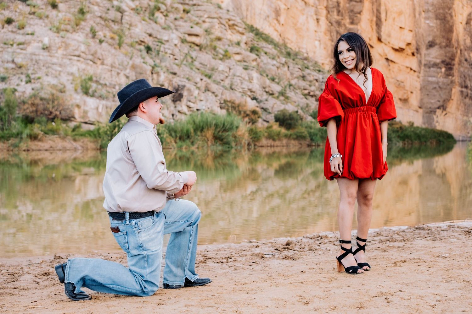 A couple in western attire shares romantic moments by a desert lake.
