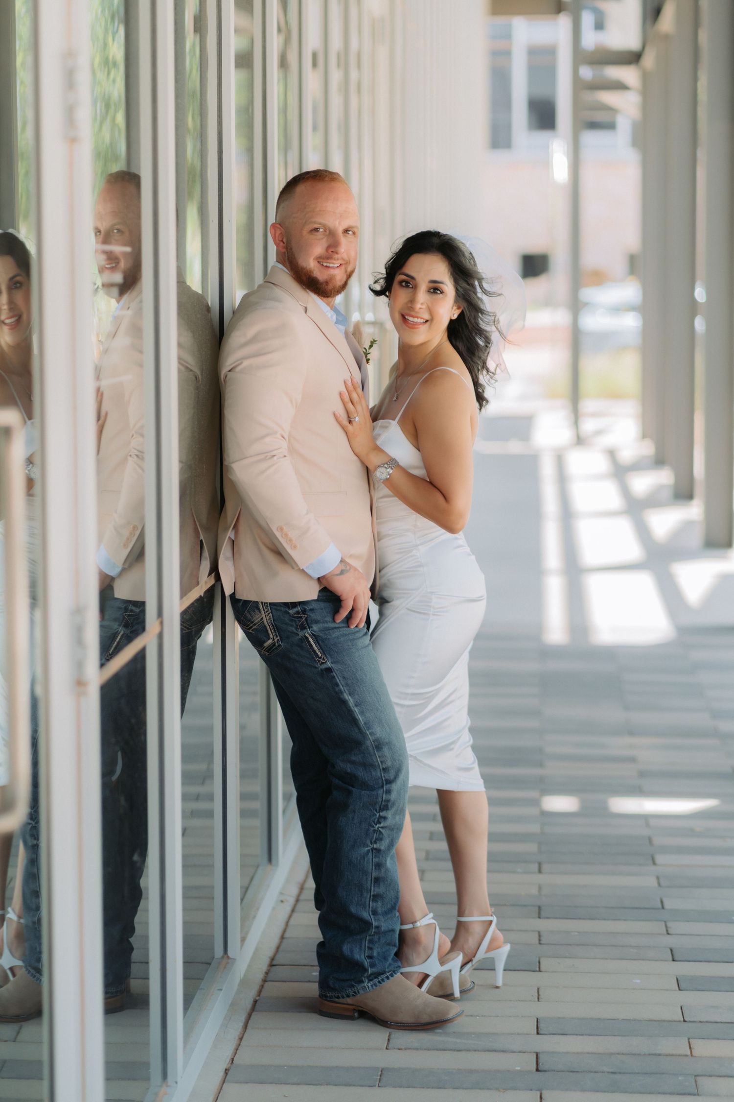 A couple in formal attire poses together in a modern hallway with reflective glass windows.