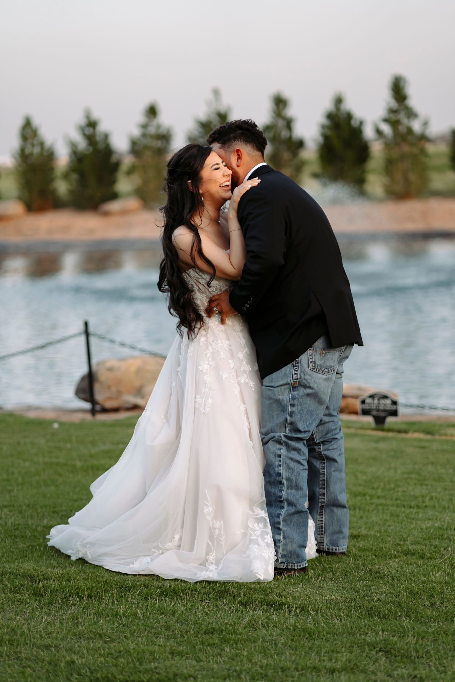 Newlyweds share an intimate moment on a grassy lawn beside a serene lake during their wedding photoshoot.