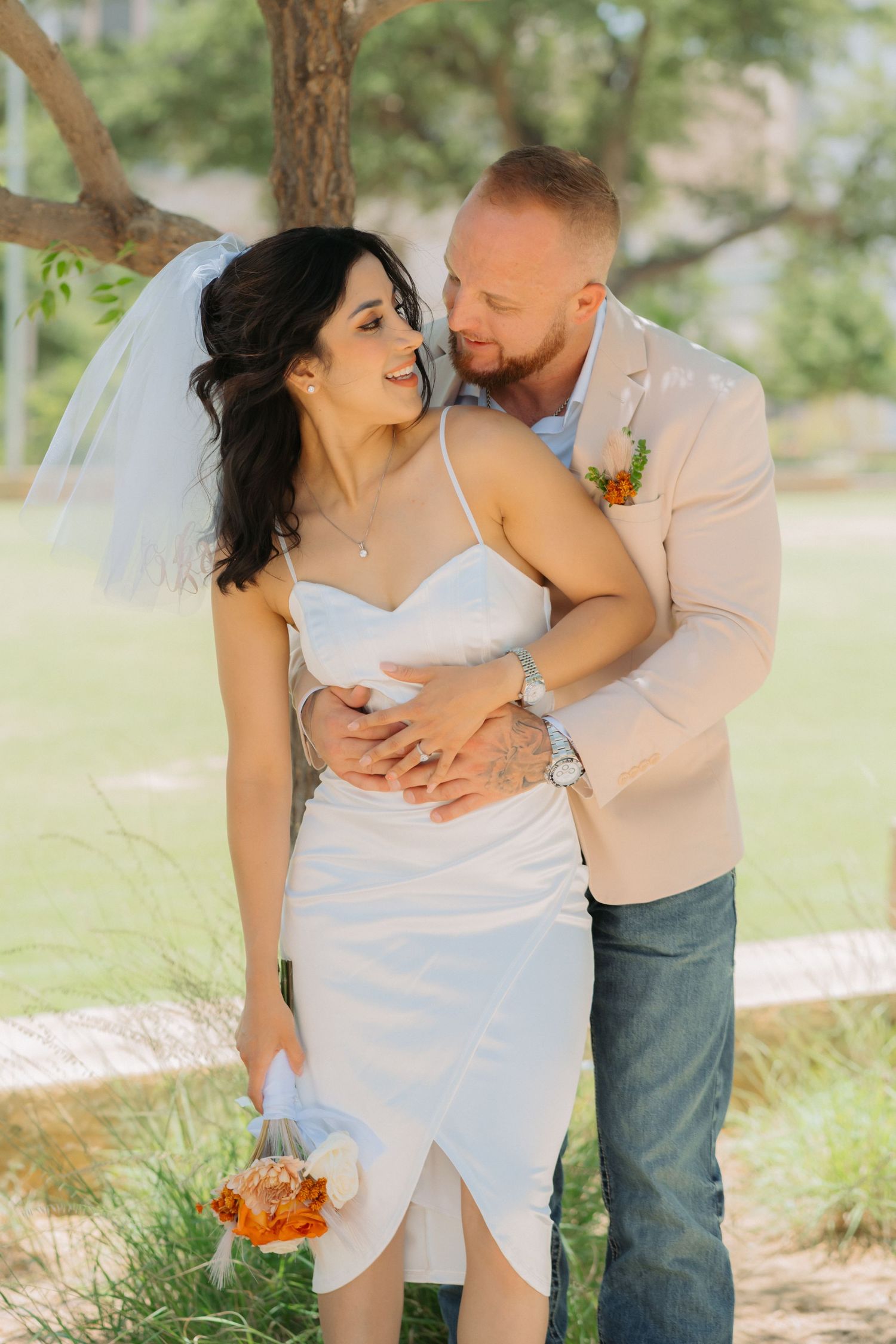 A couple embraces on their wedding day in a soft outdoor setting with greenery and natural light.