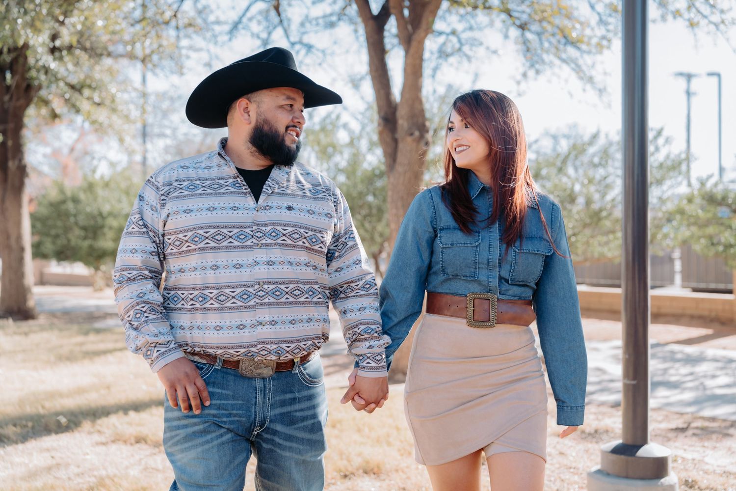 Couple in western wear smiles and poses under trees during outdoor shoot.
