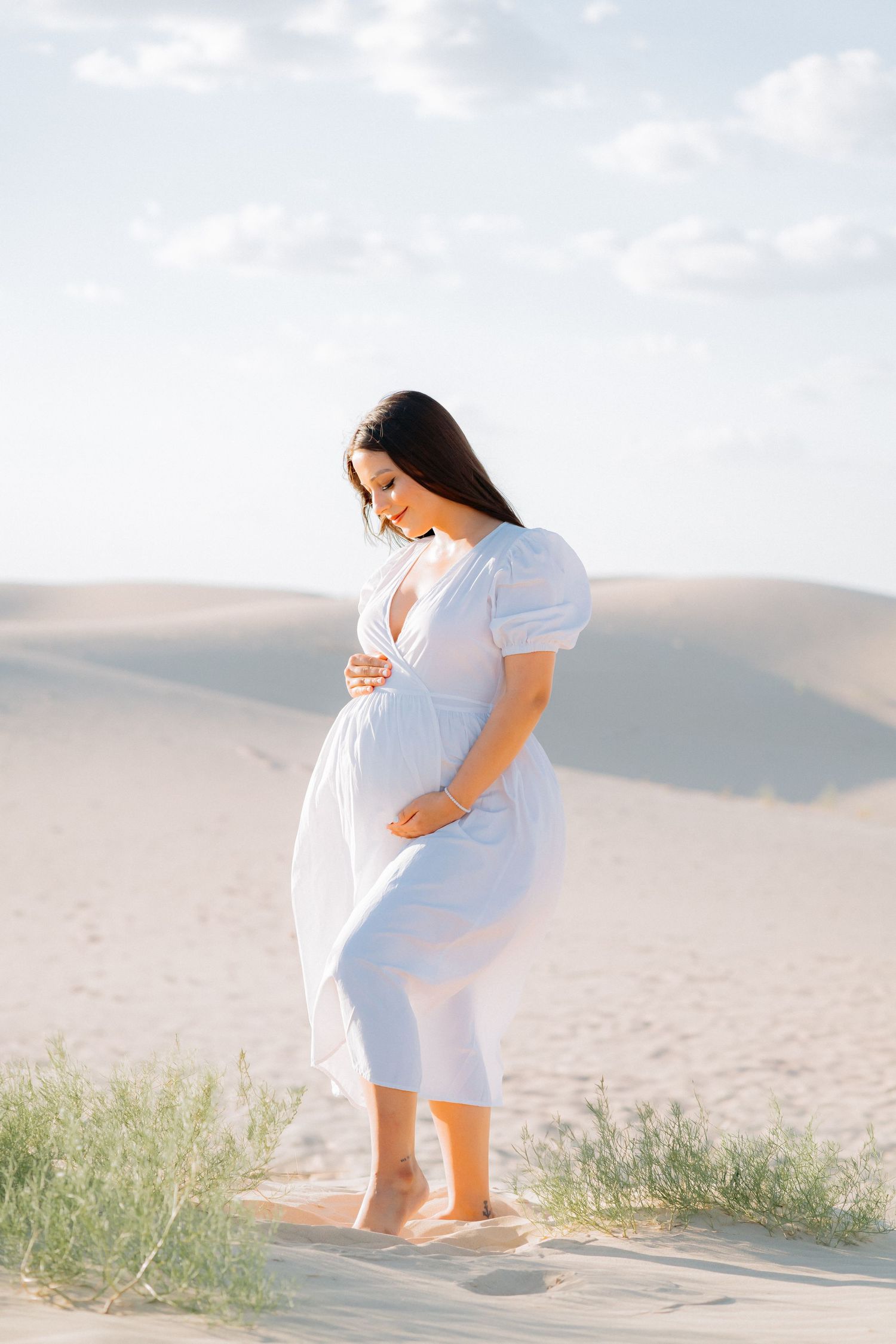 Pregnant woman in flowing white dress poses on sand dunes during golden hour.