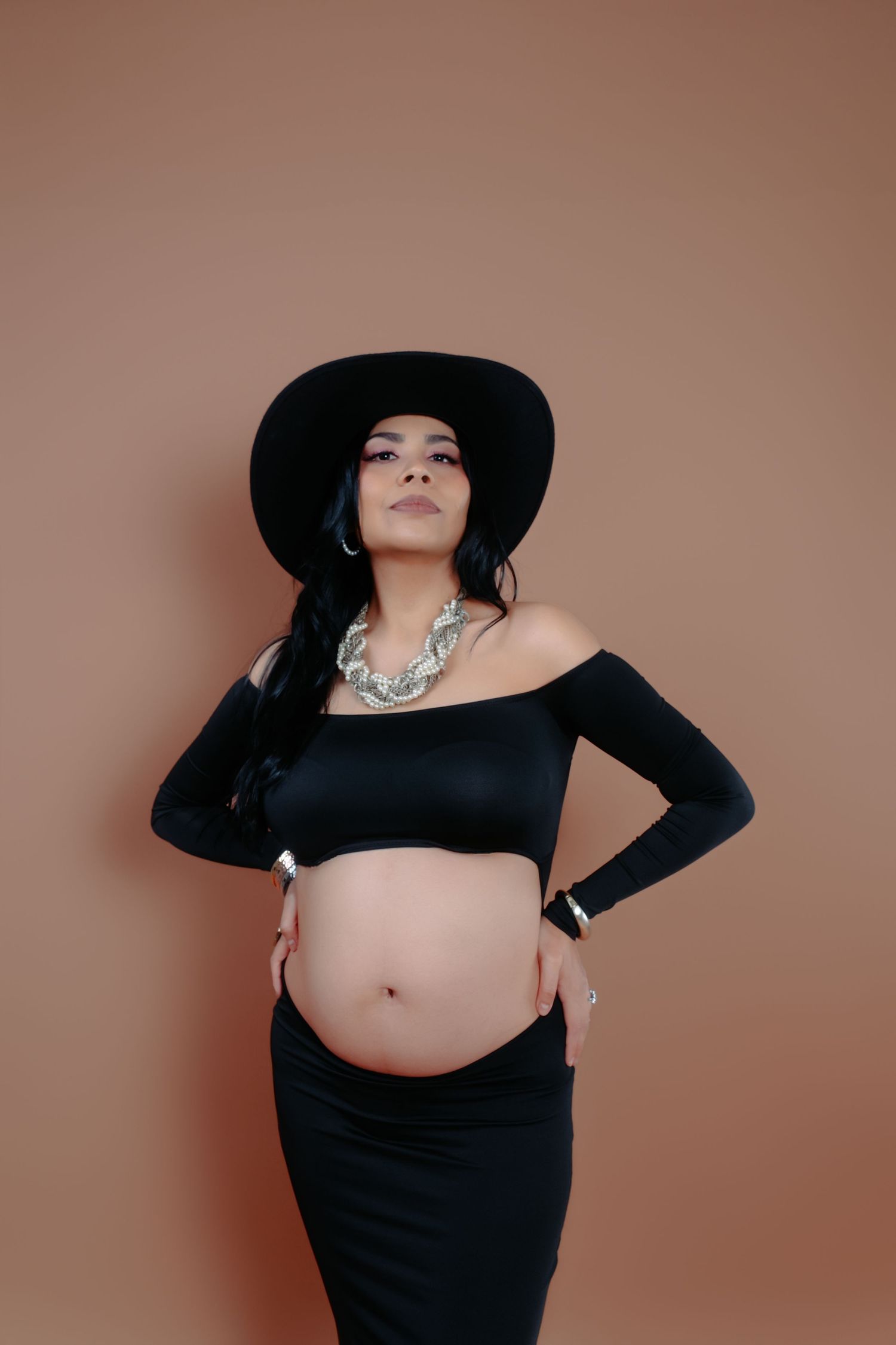 Stylish maternity portrait with woman in black crop top and hat against brown backdrop.