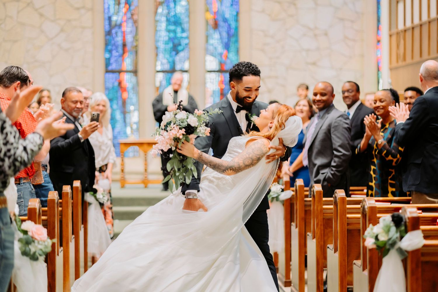 A joyful dip kiss during a wedding ceremony as guests applaud in a church with ornate decor.