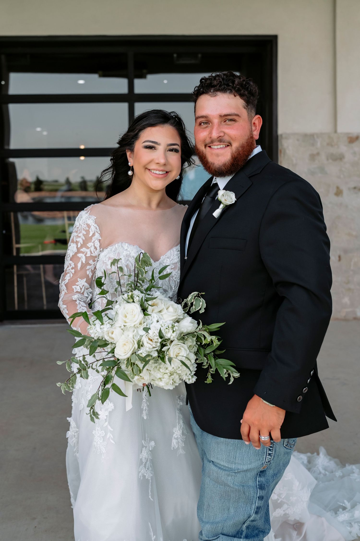 A couple in wedding attire poses together, with the bride holding a white floral bouquet with greenery.