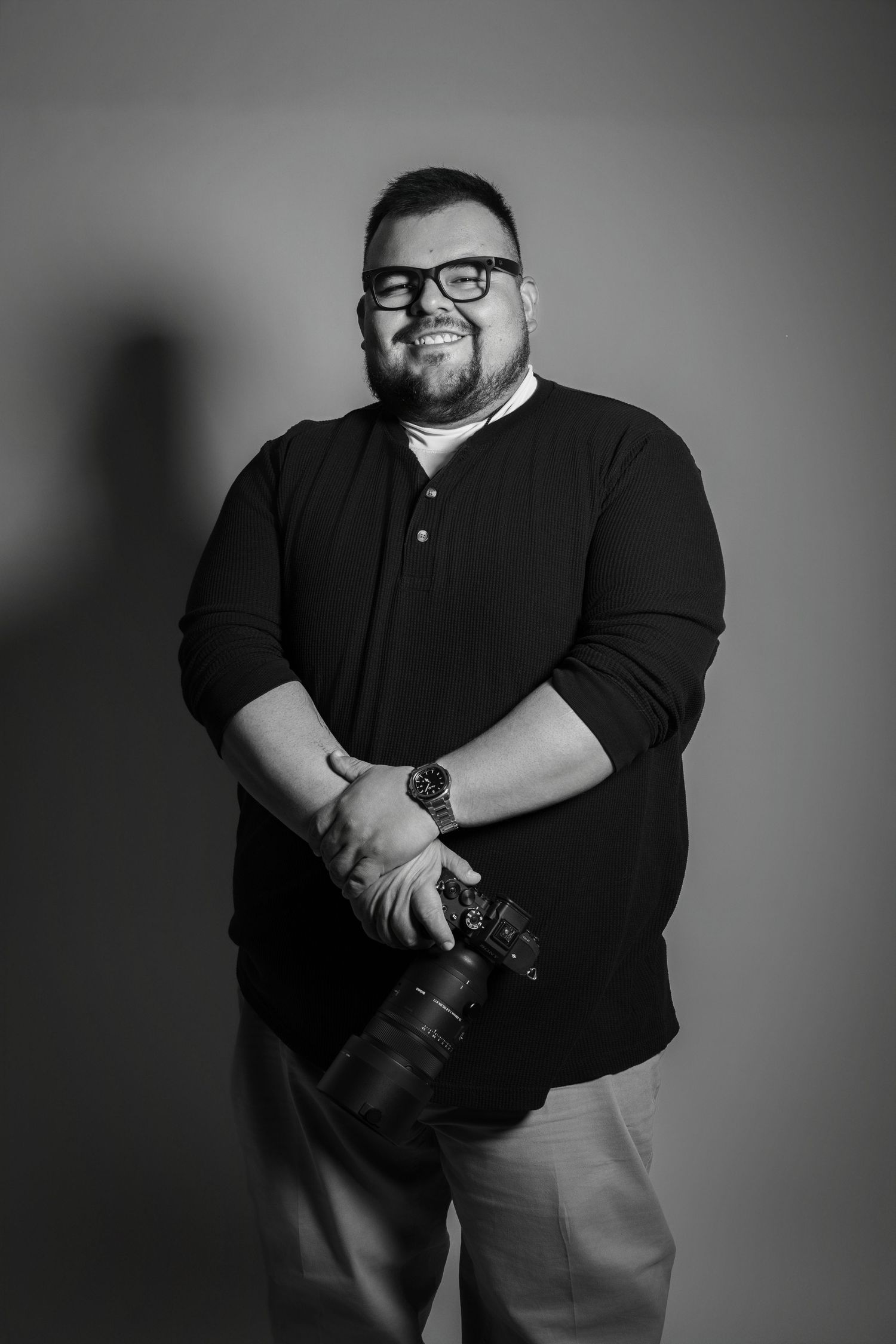 Black and white portrait of photographer holding professional camera wearing glasses and dark polo shirt against gray backdrop.