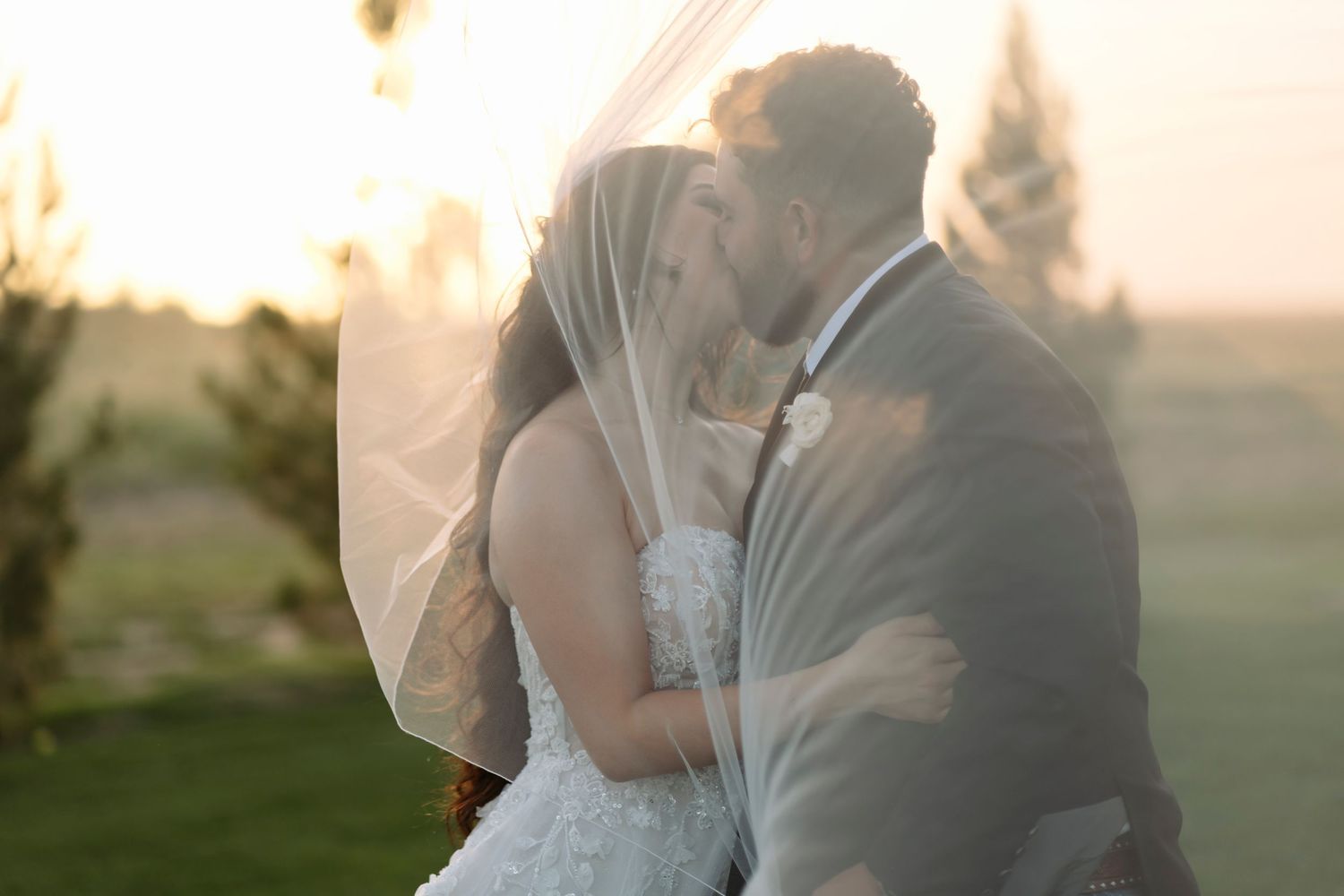 A couple in formal wedding attire shares an intimate moment during sunset in a grassy outdoor setting.