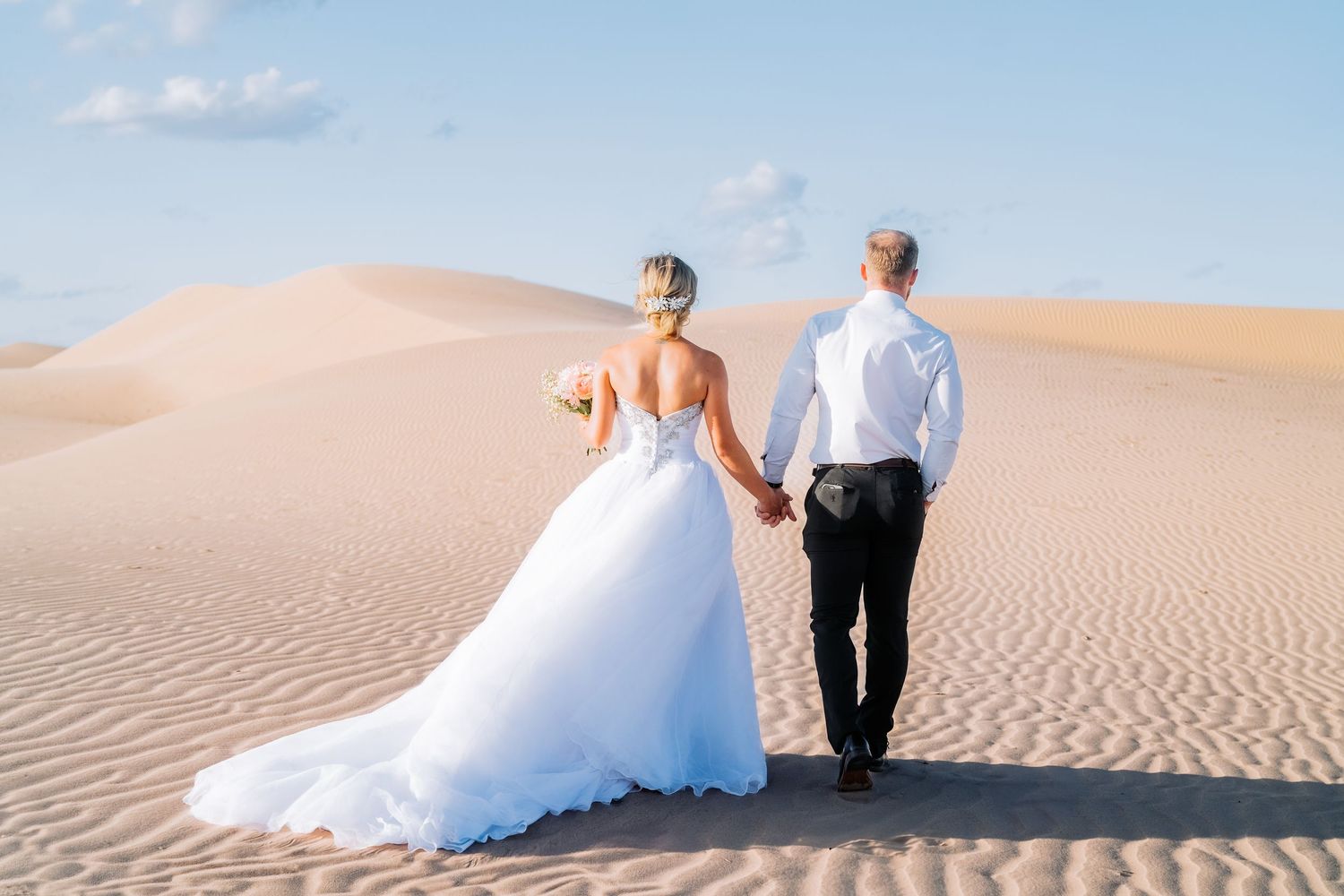 Wedding couple holding hands while walking in sand dunes under blue sky.