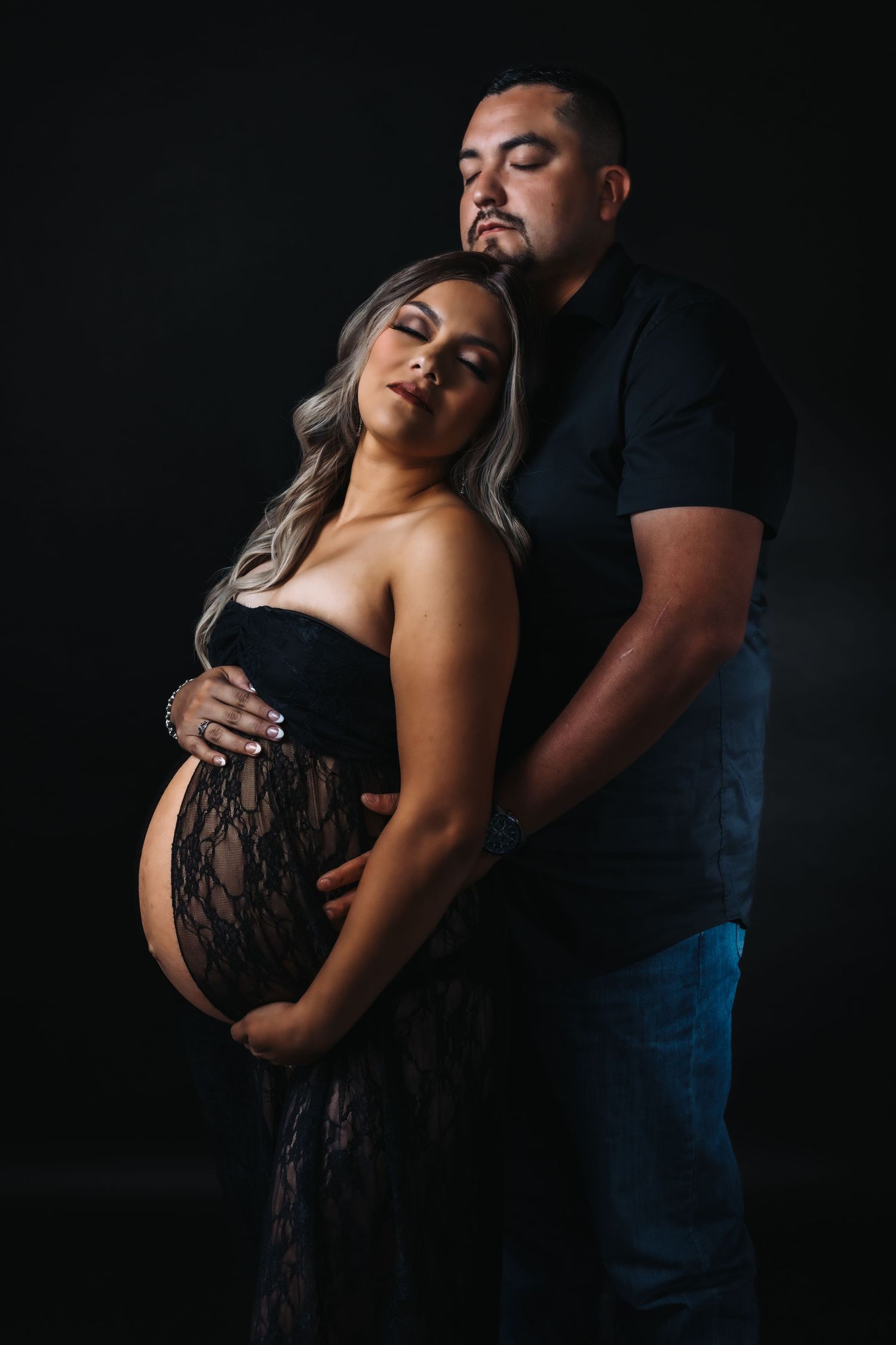 Dramatic studio maternity session featuring couple in black attire against dark background.